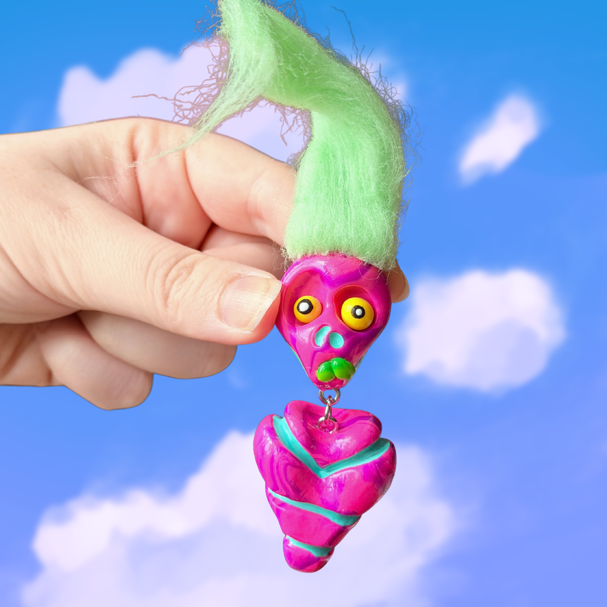Polymer clay marbled pink colored fidget that looks like a quirky skeleton with no arms or legs and light green hair. Photographed on top of a cloudy sky background.