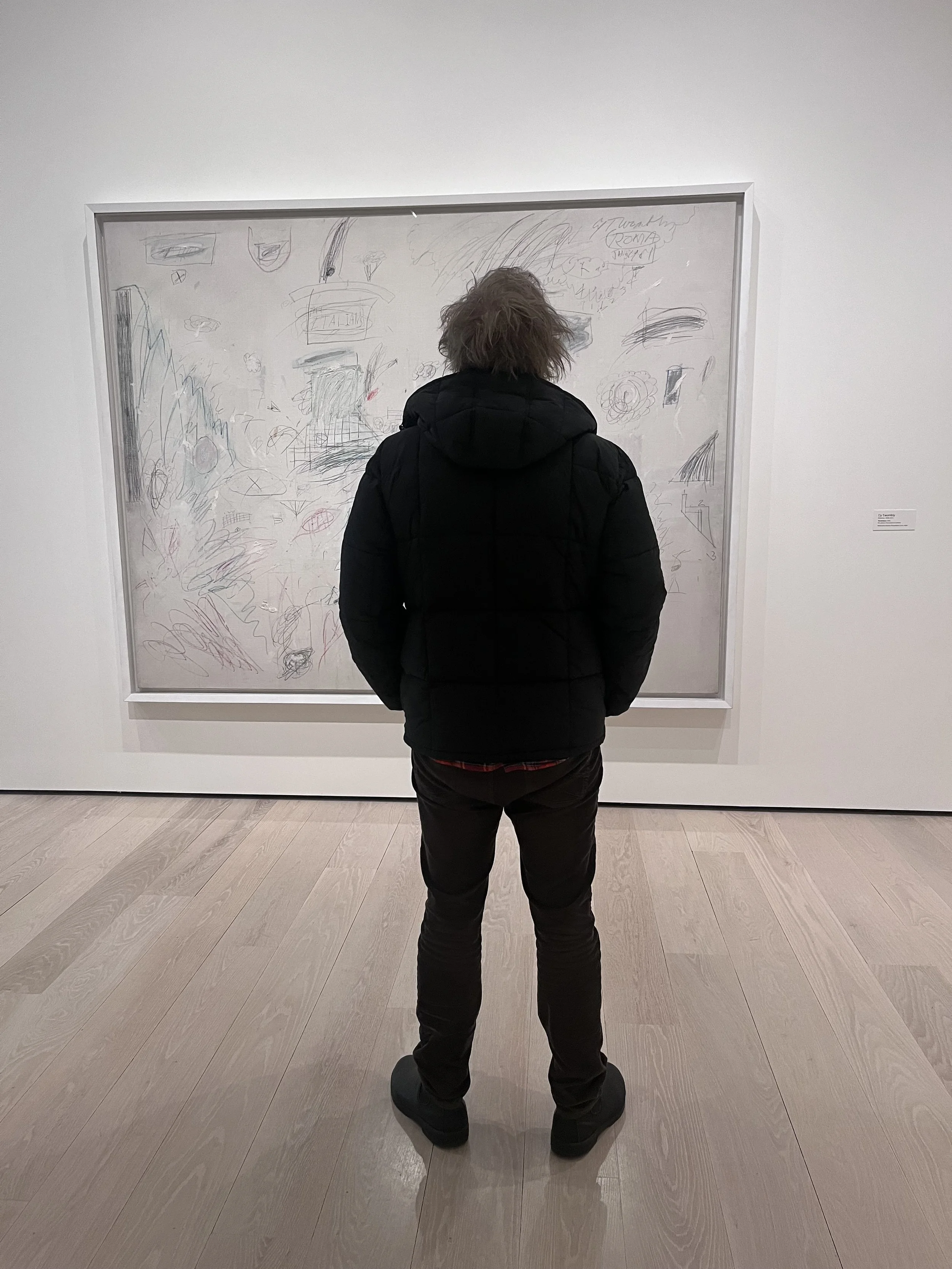 A person dressed in a black jacket and black pants, standing in front of a large painting in an art gallery.