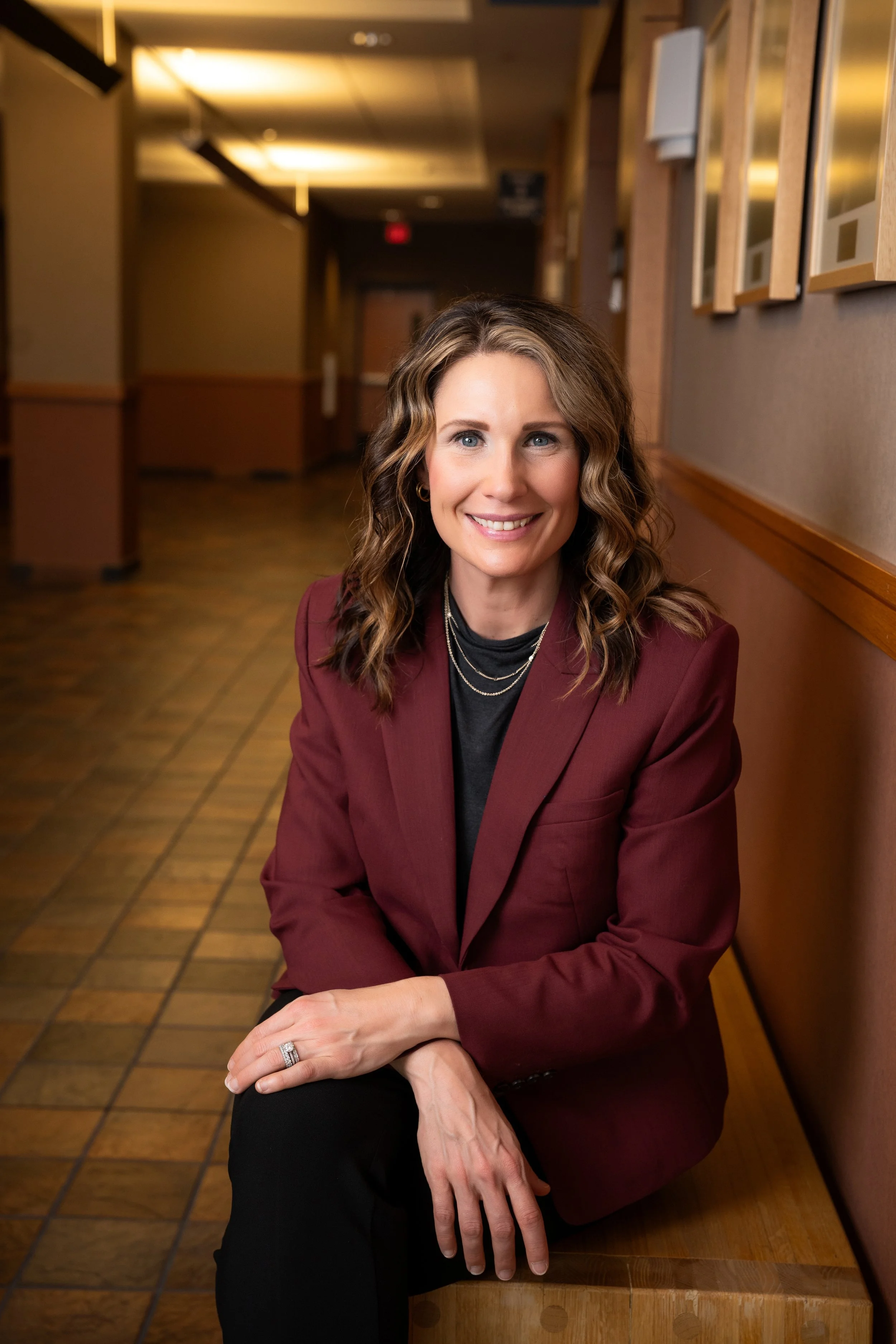 A woman with wavy brown hair, wearing a maroon blazer and black shirt, sitting at a table in a warmly lit restaurant or cafe, smiling and looking at the camera.