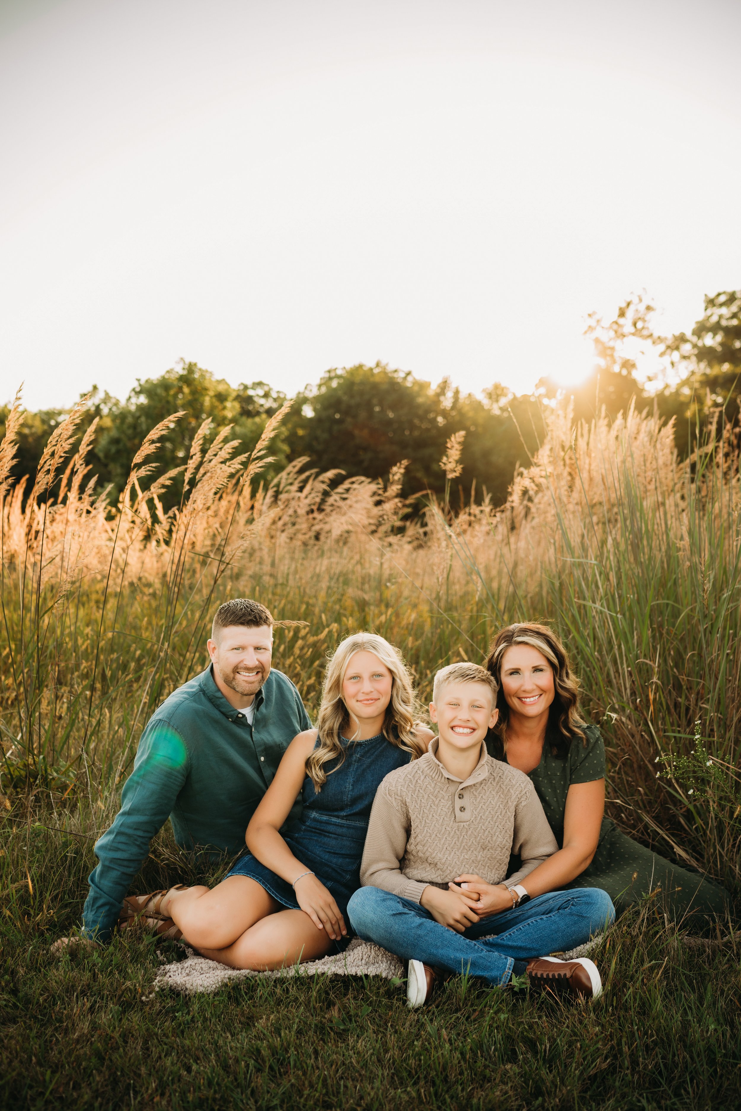 A family of five sitting together outdoors during sunset, surrounded by tall grasses and trees in the background, smiling at the camera.