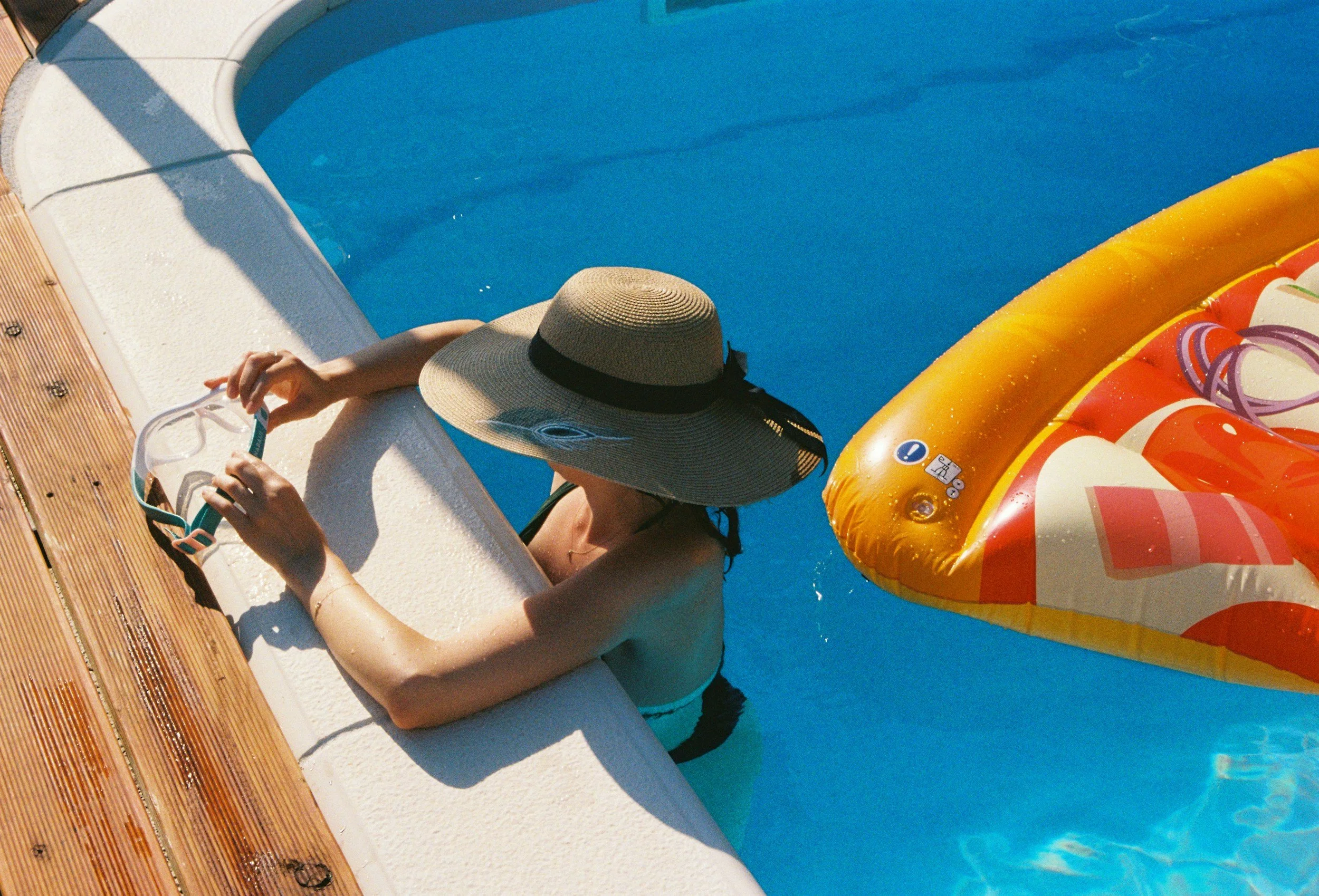 A woman wearing a large sunhat holding goggles while sitting at the edge of a swimming pool with a yellow and red inflatable raft in the water.