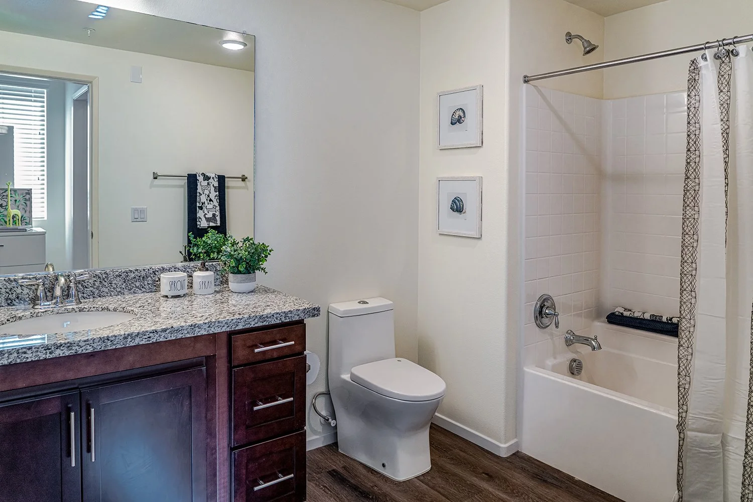 Second bathroom in Turtle Creek Apartment Home with wood-style plank flooring, large vanity mirror, granite countertops, dark wood shaker cabinets, and a shower/tub. 