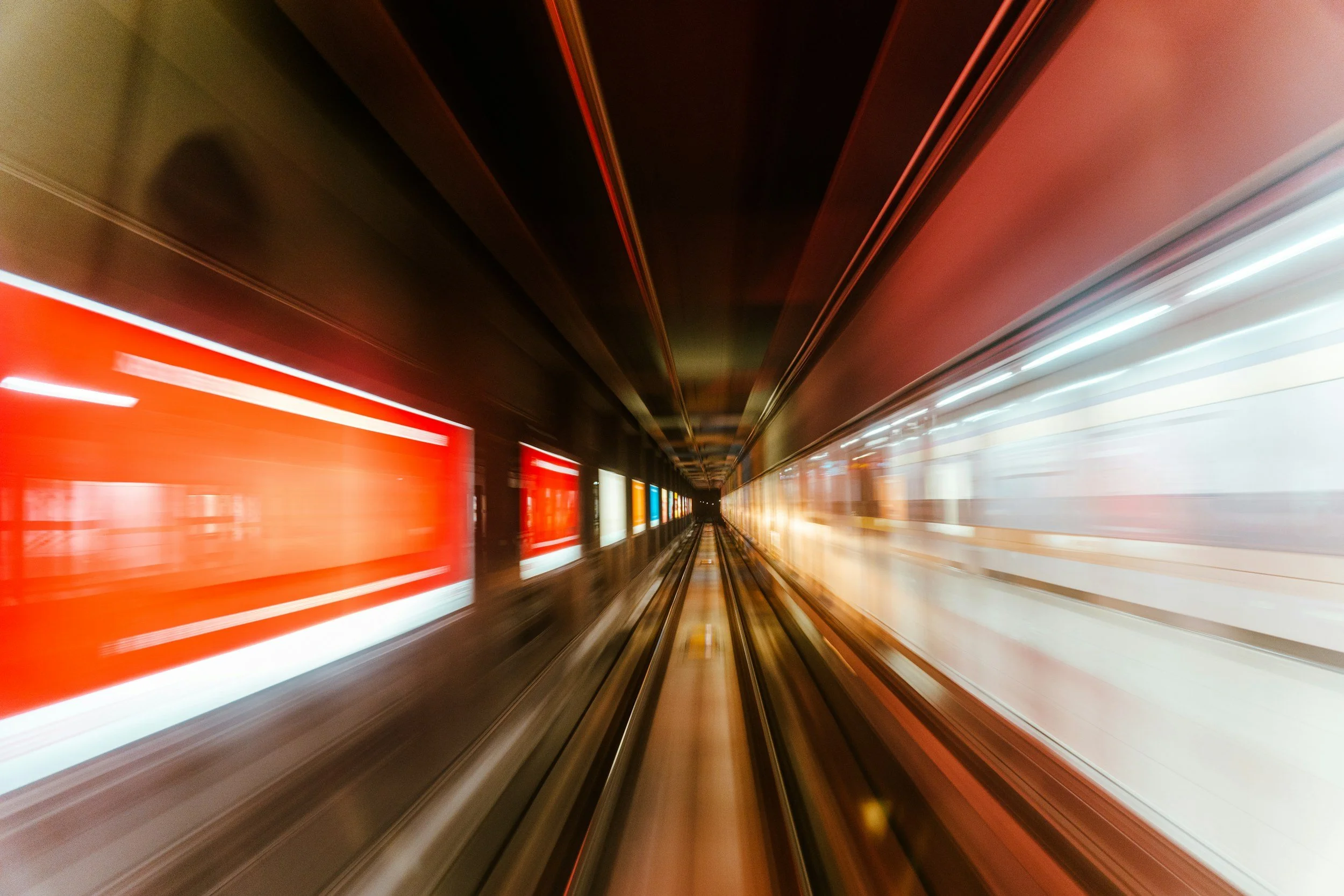 A blurred image of a train moving through an underground tunnel, with red advertisement lights on the side and streaks of white and red lights from the train.