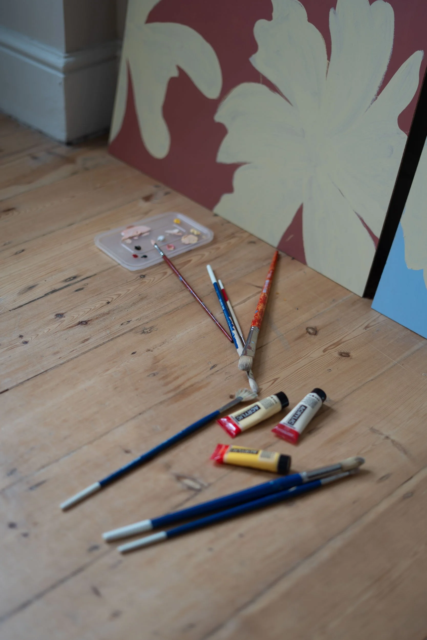 A photograph of brushes, tubes of paints and a paint palette on bare floorboards with large scale botanical paintings in the background