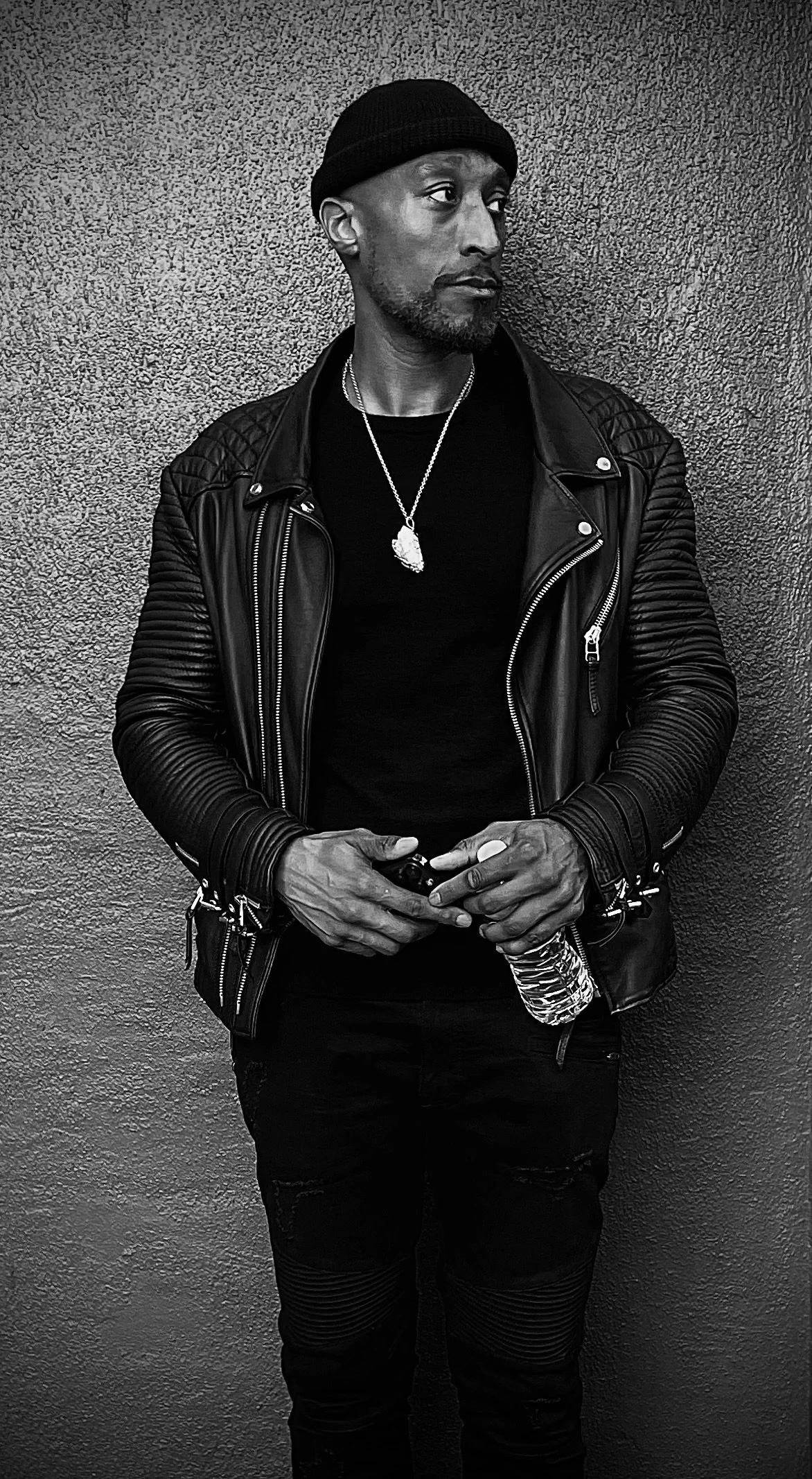 A man in a black beanie, leather jacket, black shirt, holding a water bottle, standing against a textured wall.