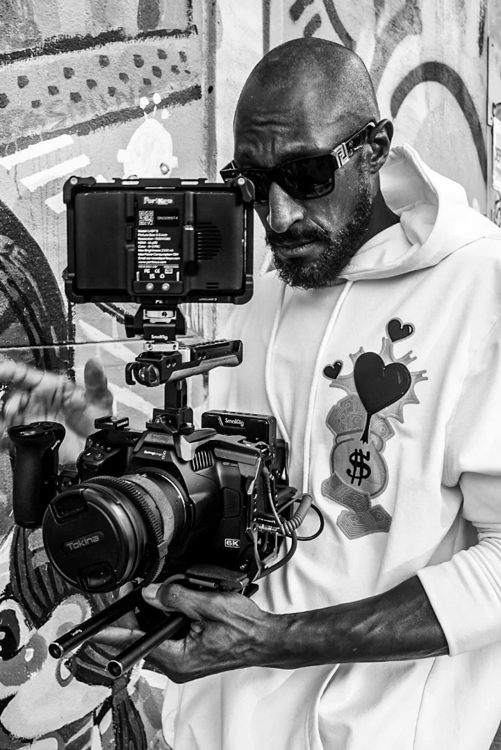 Man holding a professional camera setup, including a monitor, in front of graffiti wall, wearing sunglasses and a hoodie with graphic design.