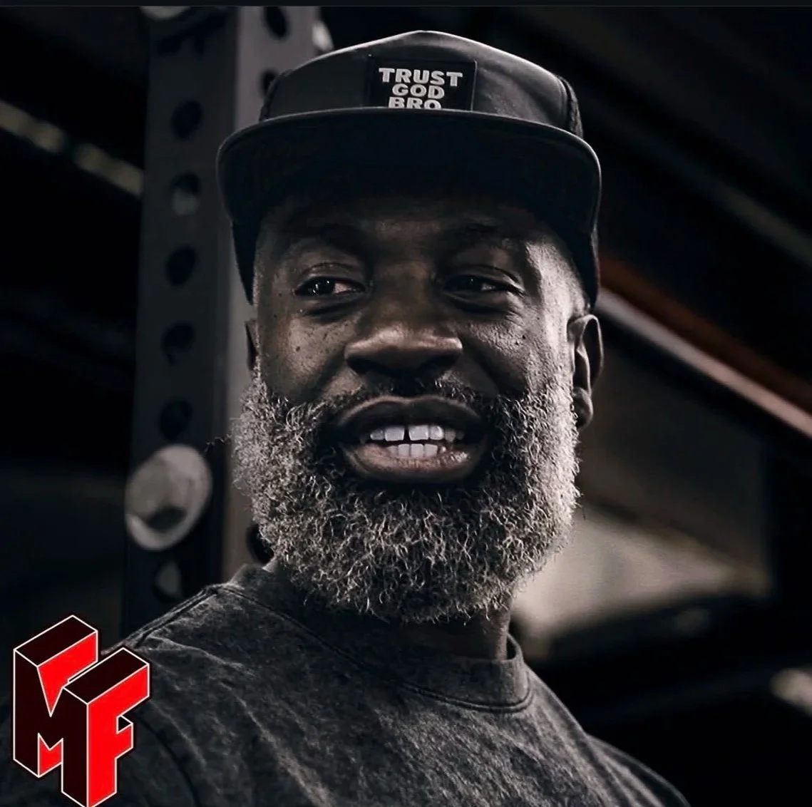 A man with a gray beard and mustache wearing a black cap that says 'Trust God Bro' and a black t-shirt, standing indoors in front of a metal support beam.