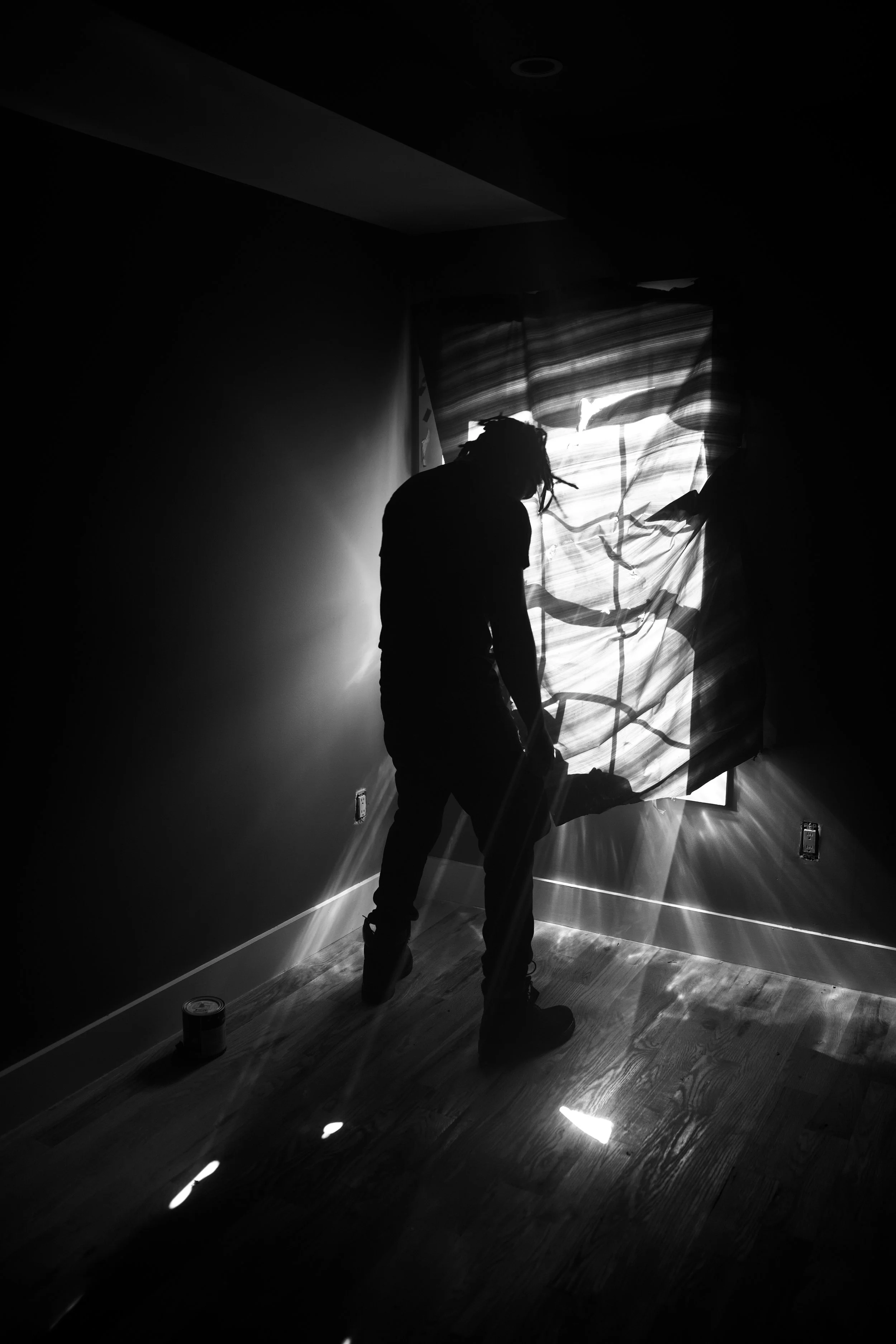 Silhouette of a person with dreadlocks working with a large abstract or artistic fabric backdrop in a dimly lit room with sunlight streaming through.