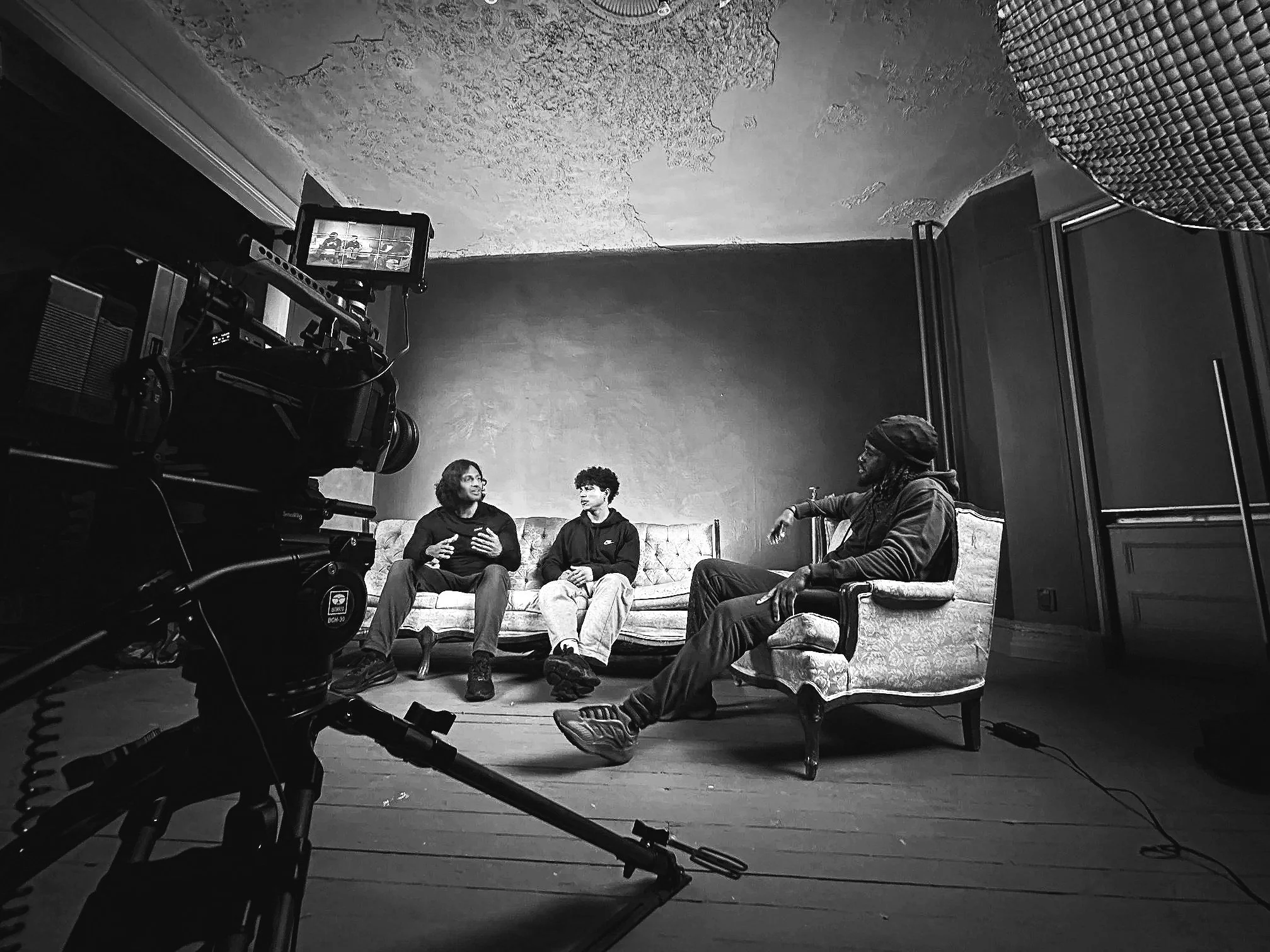 Black and white photo of three people sitting on a sofa and talking, with a camera set up in the foreground capturing their conversation, in a room with a peeling paint ceiling and a plain wall in the background.