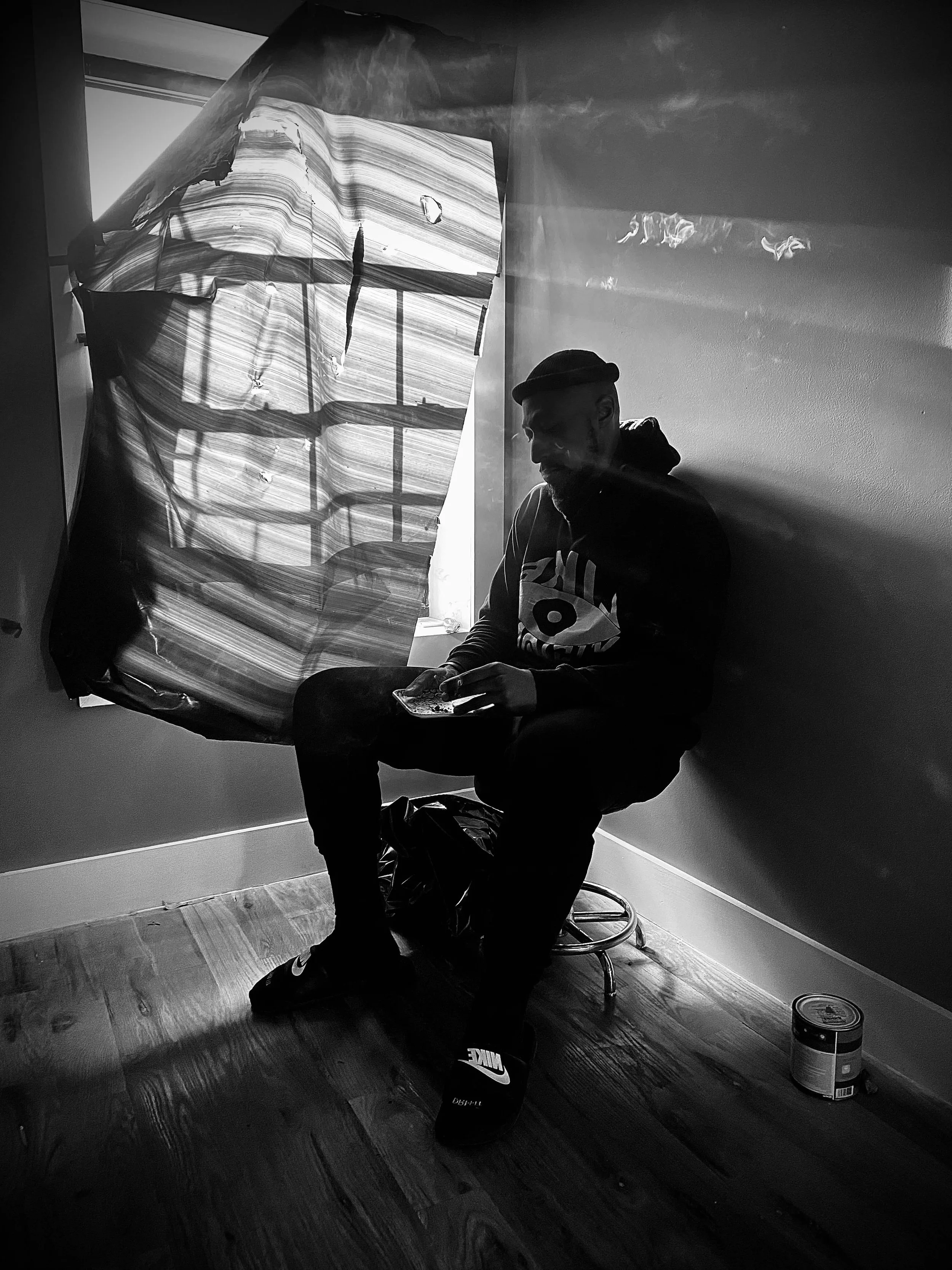 A young man sitting on a stool in a room with sunlight streaming through a window. He is wearing a beanie, hoodie, and sneakers, looking down at his phone. There is a torn fabric curtain or sheet hanging behind him, and a small can of paint or similar container on the floor nearby.