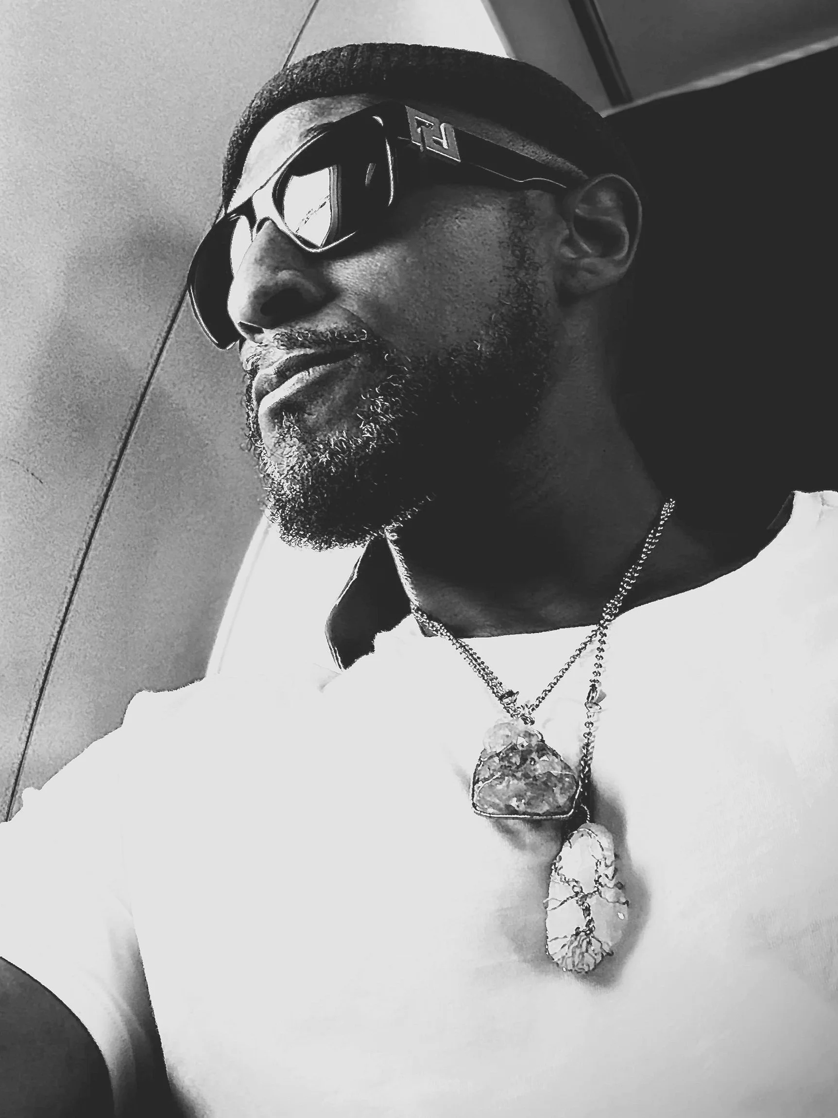 A person with a beard wearing sunglasses, a hat, and layered necklaces, looking to the side in black and white.