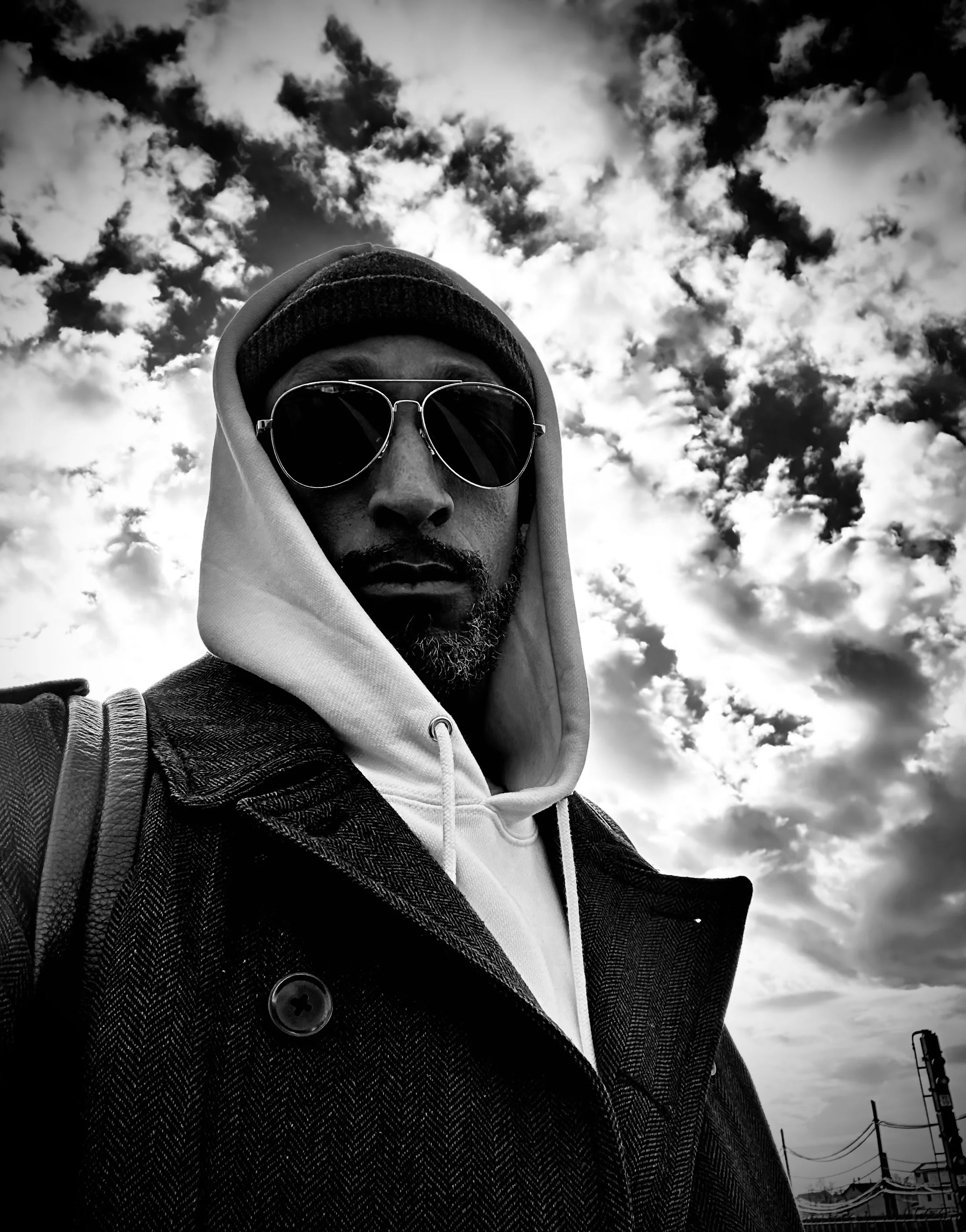 A man wearing sunglasses, a hoodie, and a coat, taking a selfie outdoors with a cloudy sky overhead.