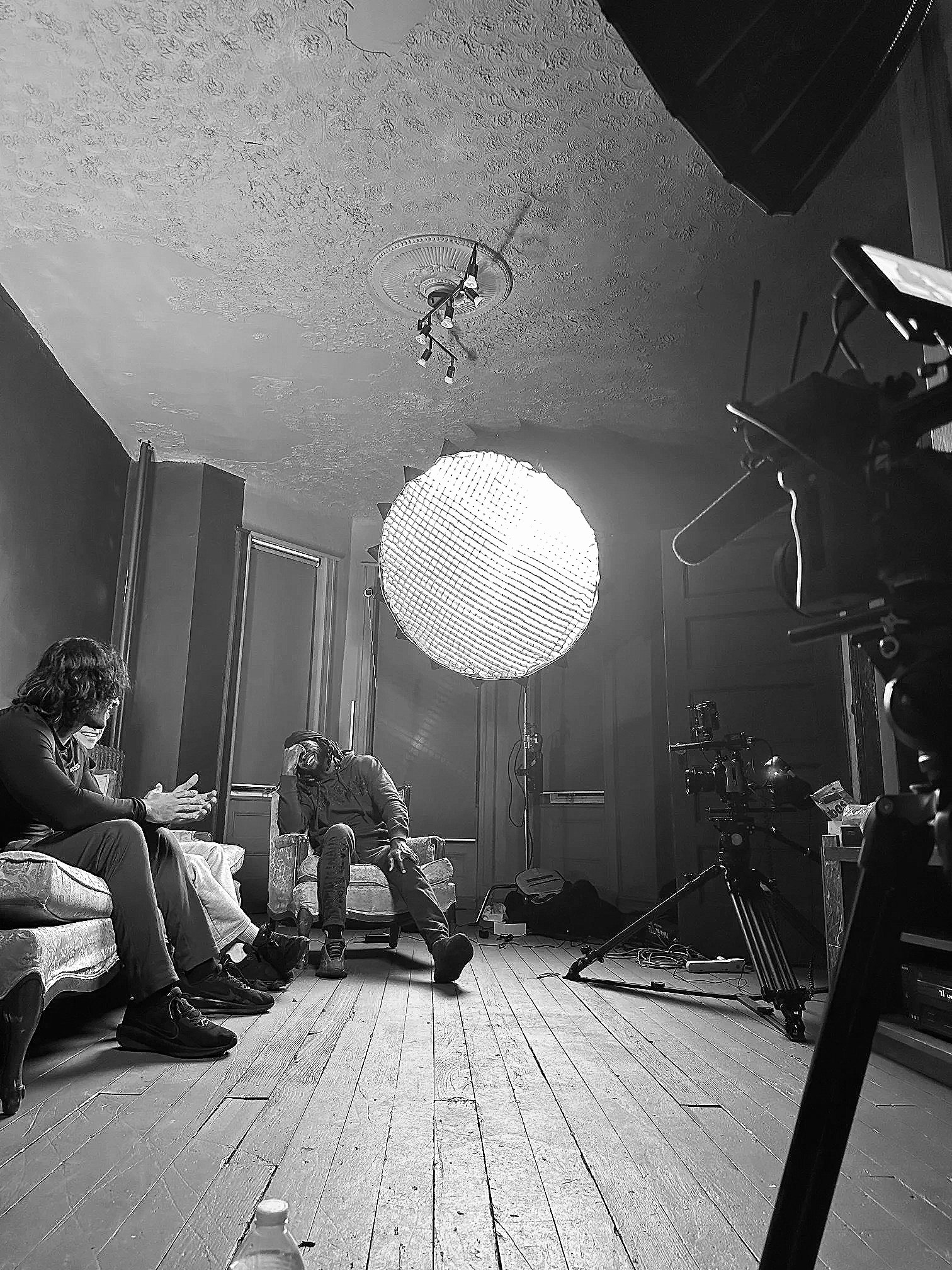 A behind-the-scenes photo of a photo shoot or interview setup in a room with a textured ceiling, wooden floor, and paneling. There are three people seated on a couch and a chair having a discussion, with studio lighting, a camera, and a large umbrella reflector visible.