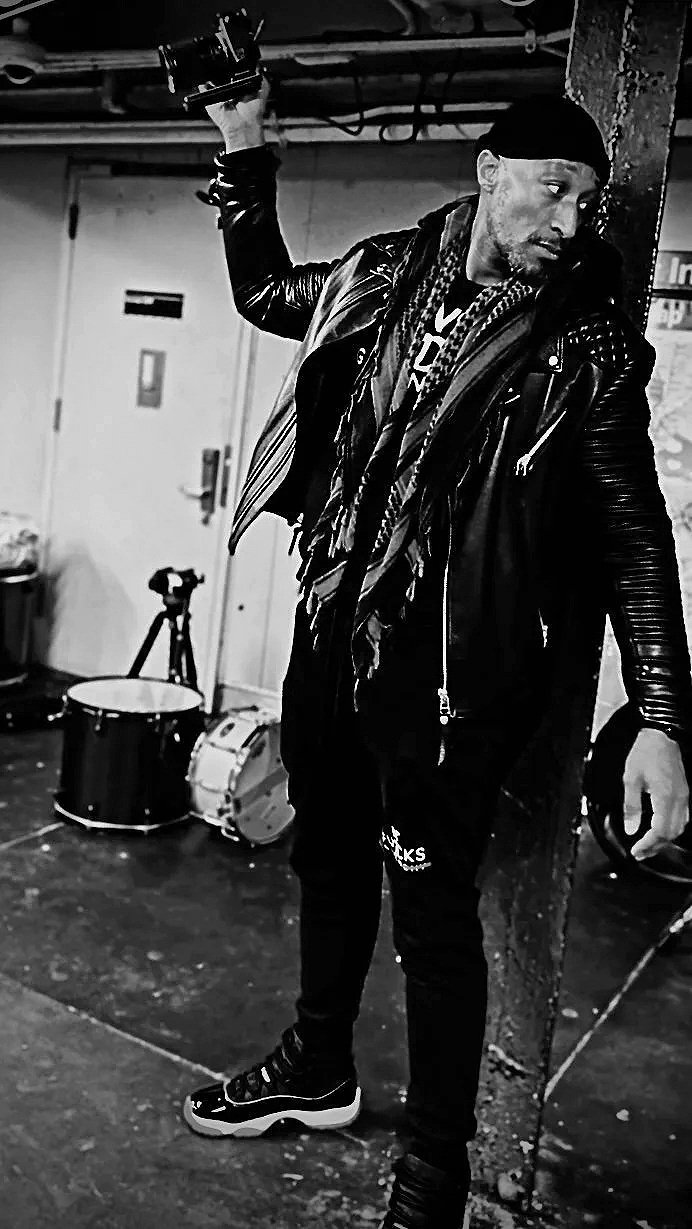 A man in a black leather jacket and black beanie holding a dumbbell in a music practice room with drums and a microphone in the background, black and white photo.