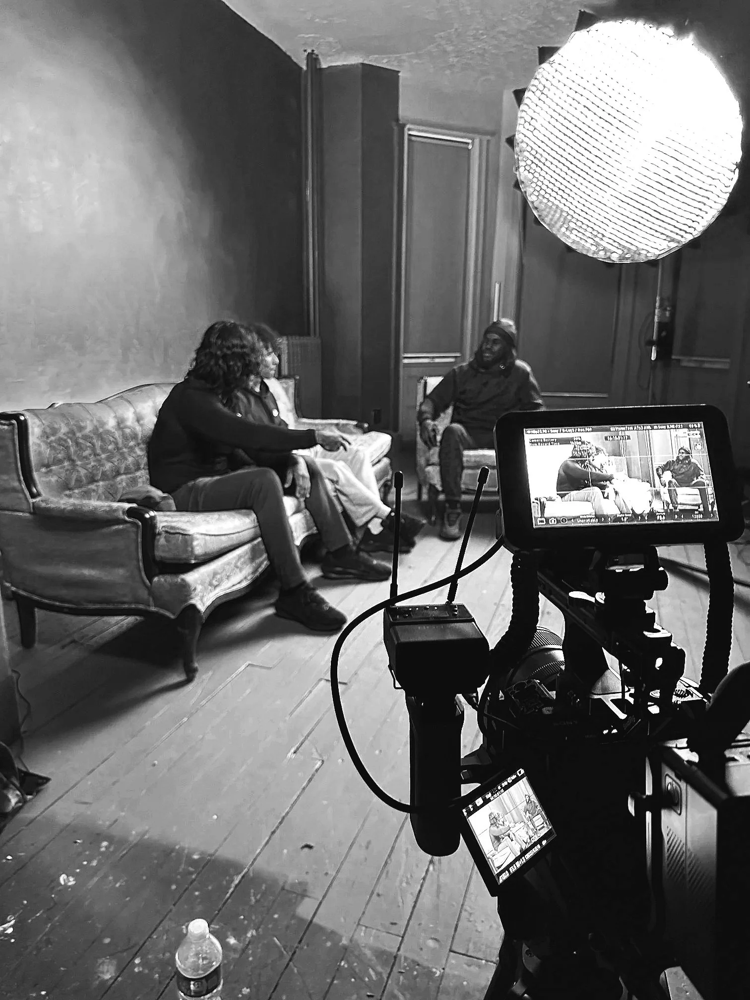 Black and white photo of a film set with three seated people in conversation, a camera on a tripod, and professional lighting equipment in a room with wooden floors and plain walls.
