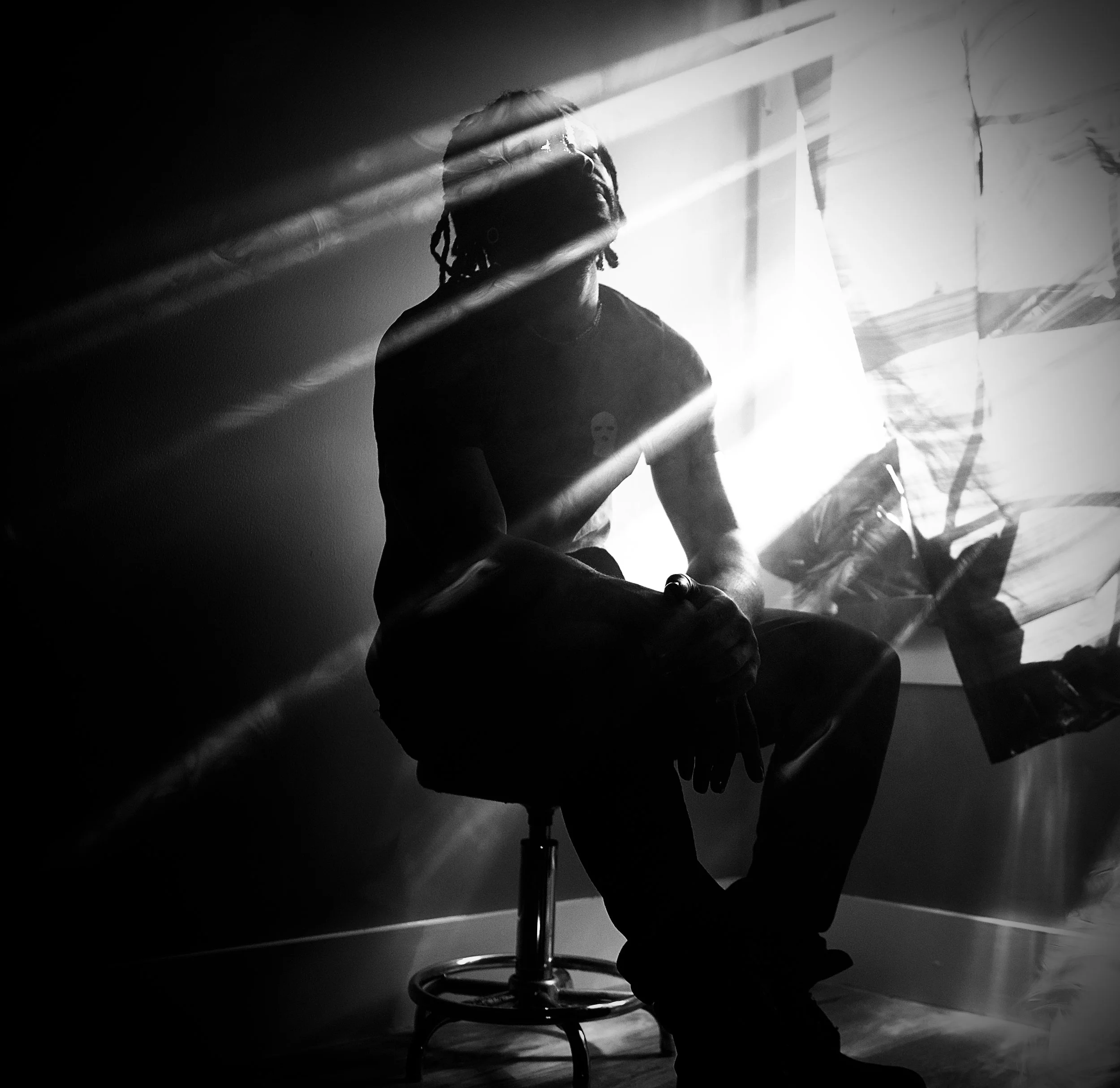 Black and white photo of a person with dreadlocks sitting next to a window with blinds, sunlight streaming through creating diagonal lines across the scene.