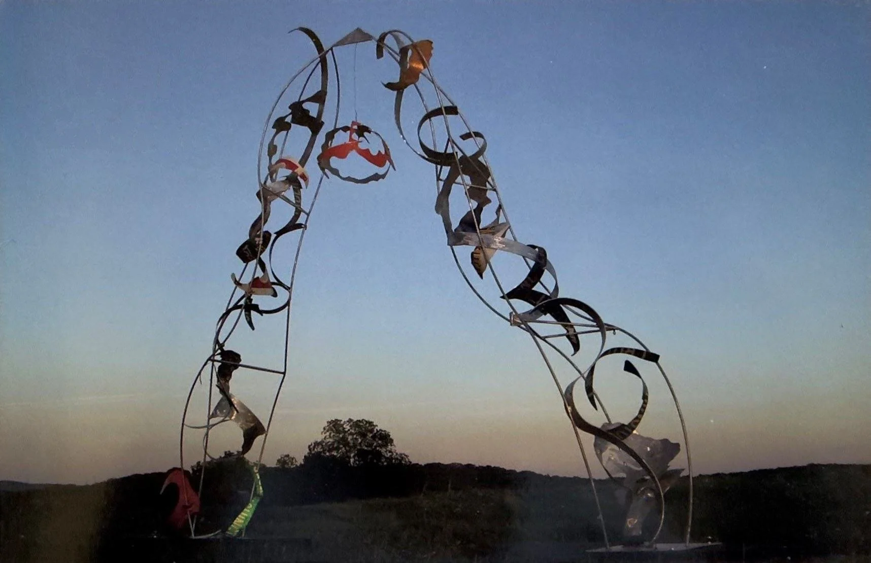Abstract metal sculpture of looping ribbons and colorful fragments silhouetted against a blue evening sky