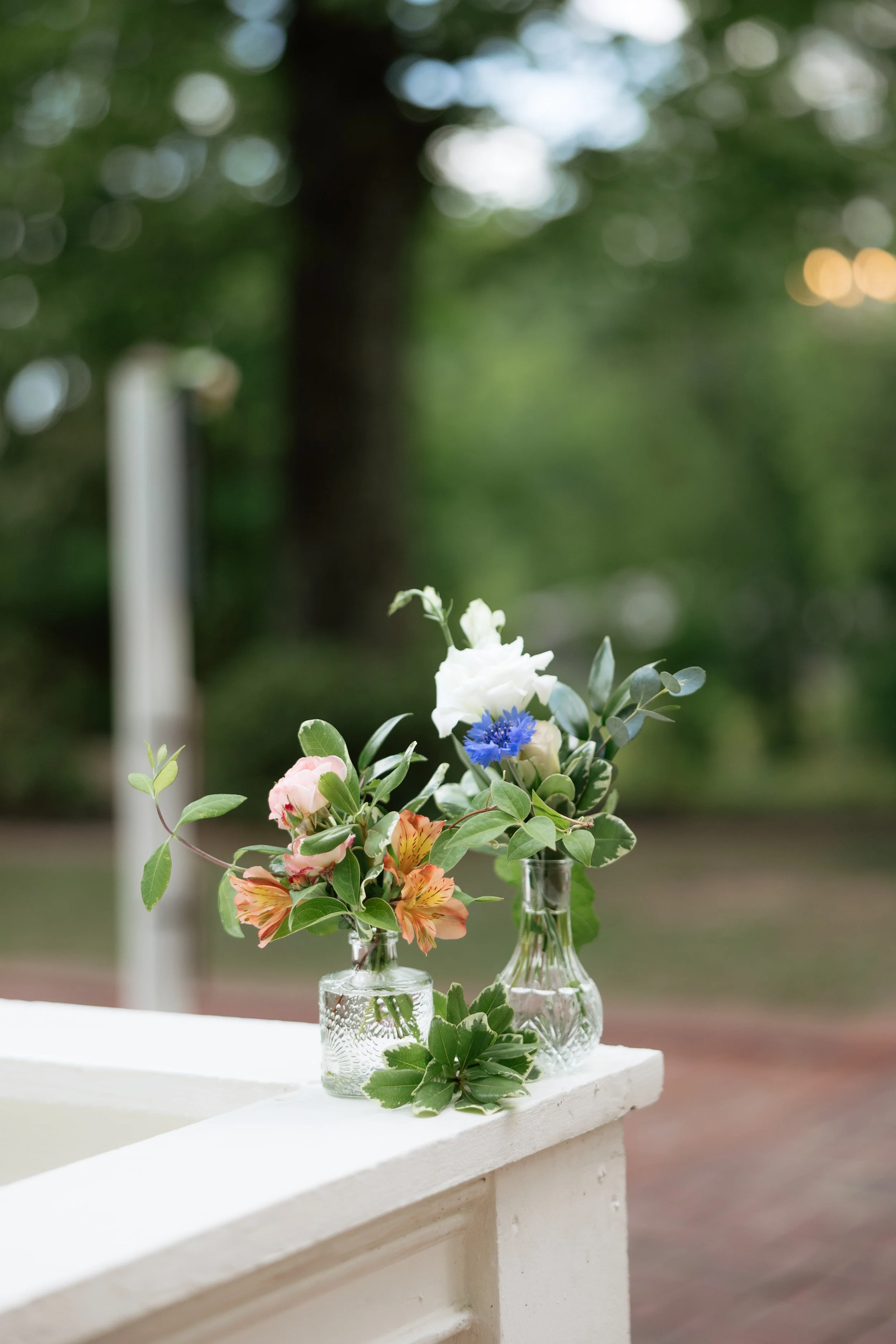 Flower arrangement with pink, white, and blue flowers in glass vases on a white table, outdoors with blurred green trees in the background.