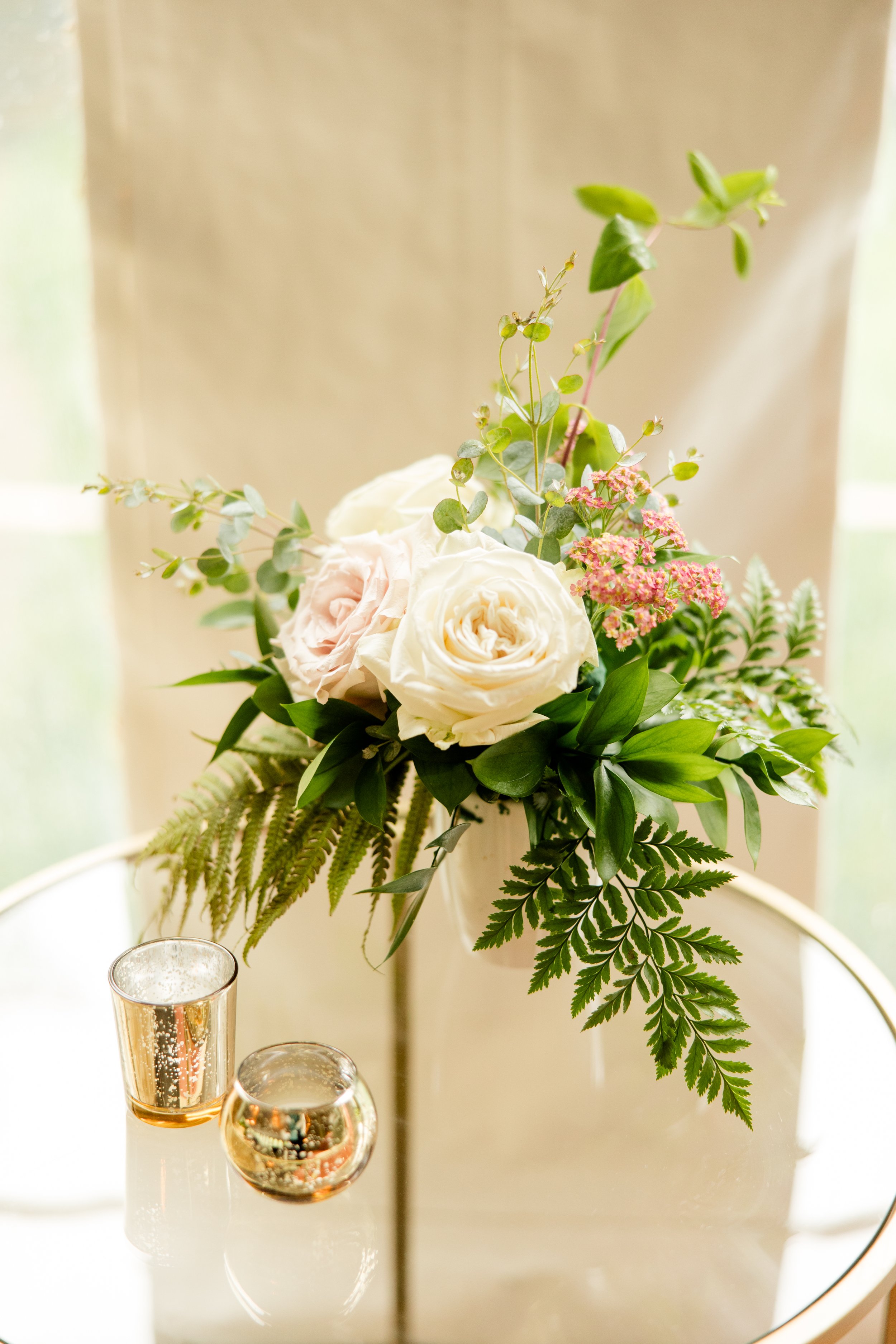 A floral arrangement with white roses, pink flowers, and various green leaves and foliage, placed on a reflective surface with two small glasses nearby._soft natural light illuminates the scene._
