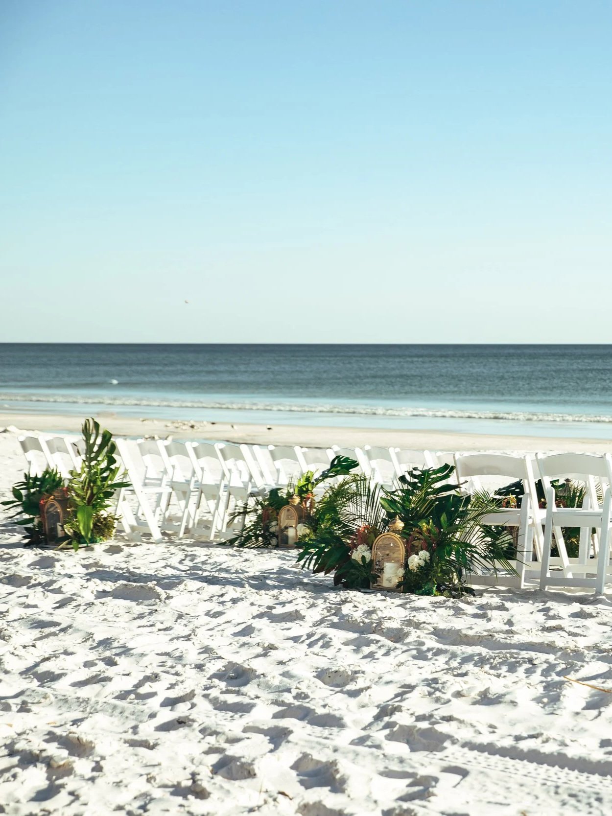 Seaside wedding setup with white chairs and floral arrangements on sandy beach, overlooking the ocean with a clear blue sky.