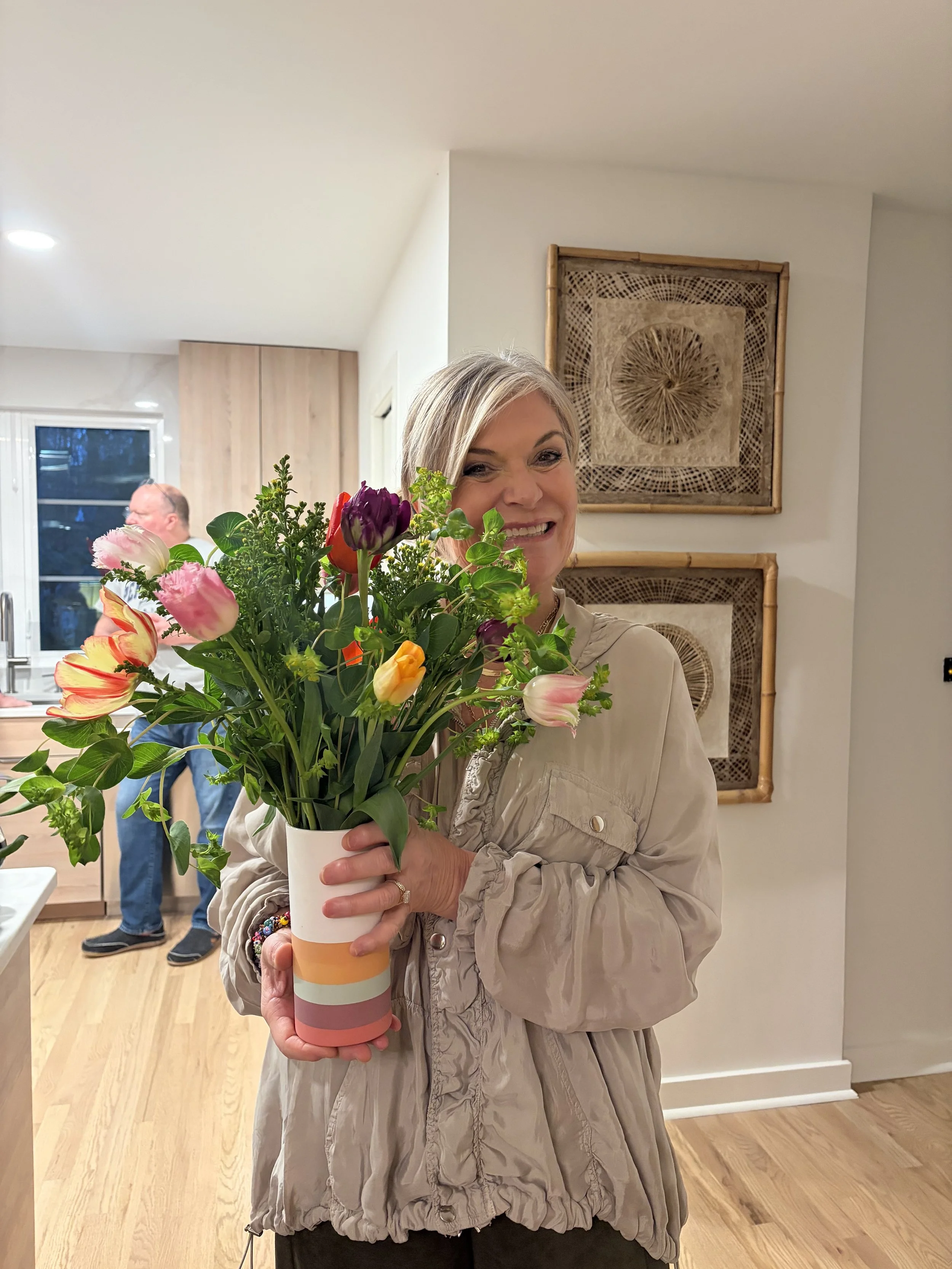 A woman smiling and holding a colorful bouquet of flowers in a white cup. In the background, a man and a child are near a window in a home interior with wooden flooring and artwork on the wall.