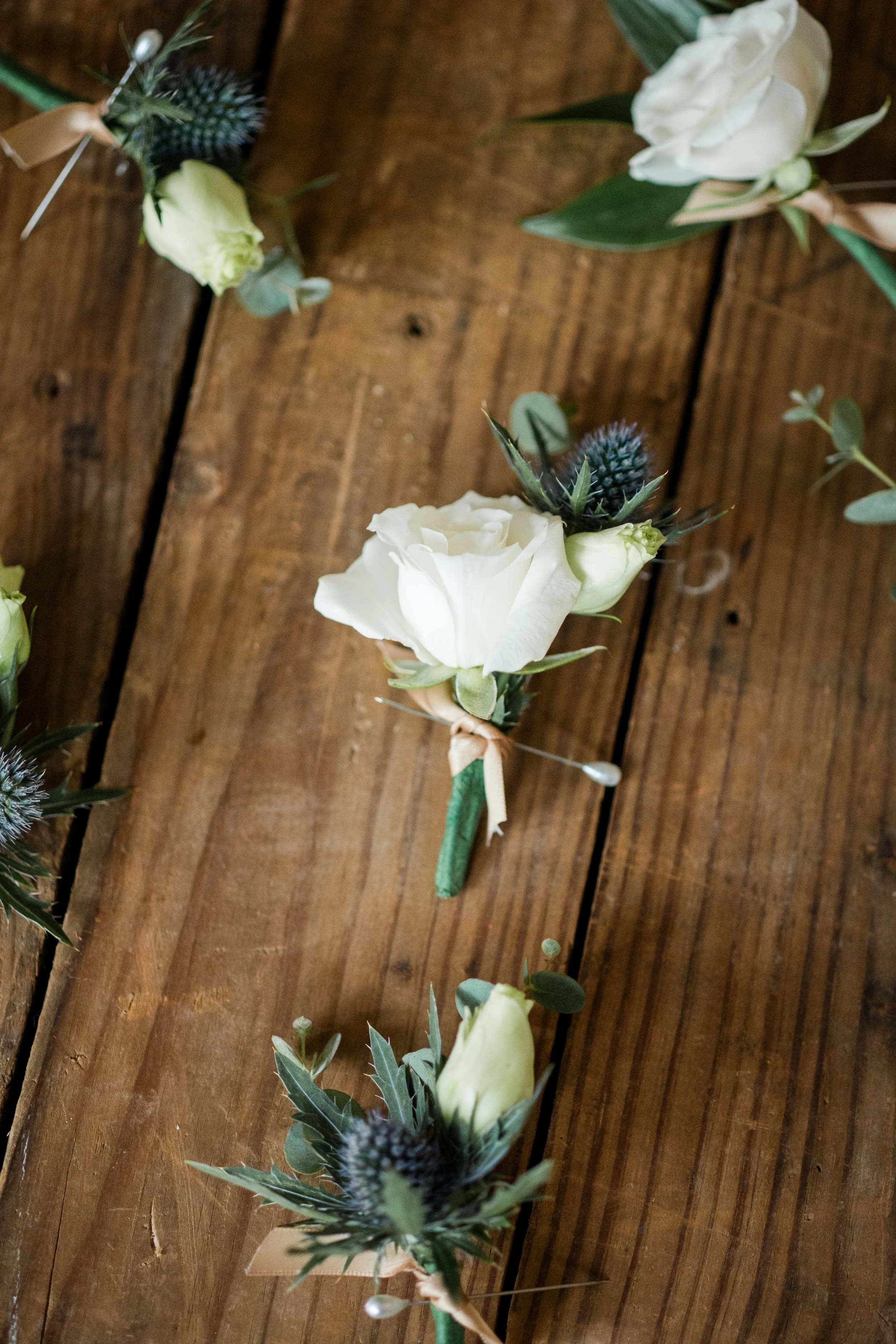 A floral boutonniere with white rose, blue thistle, and greenery pinned to a wooden surface.