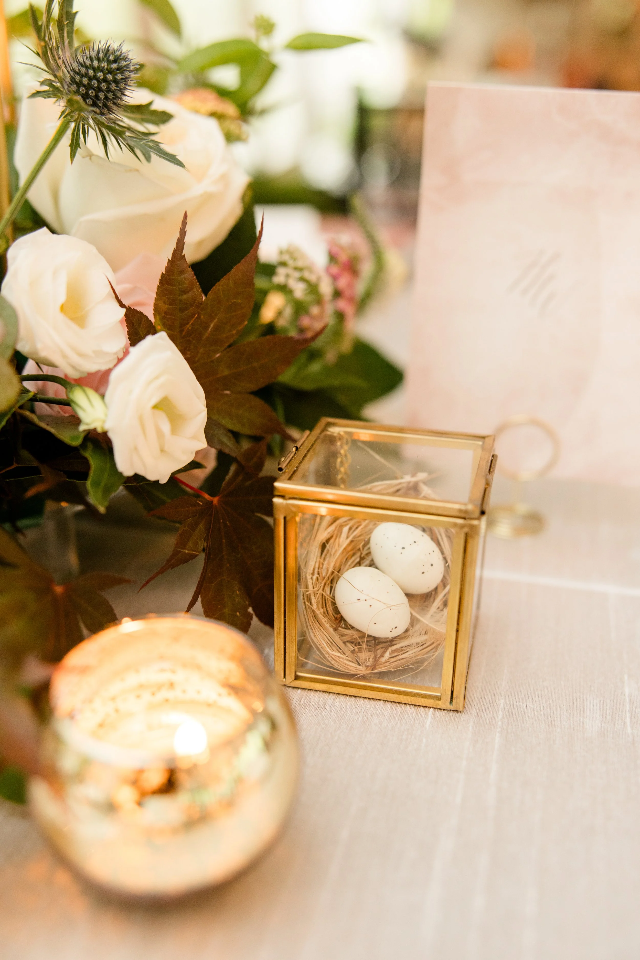 A decorative arrangement featuring white roses, maroon leaves, and other greenery, with a small glass box containing three speckled eggs and a candle in a glass holder nearby.