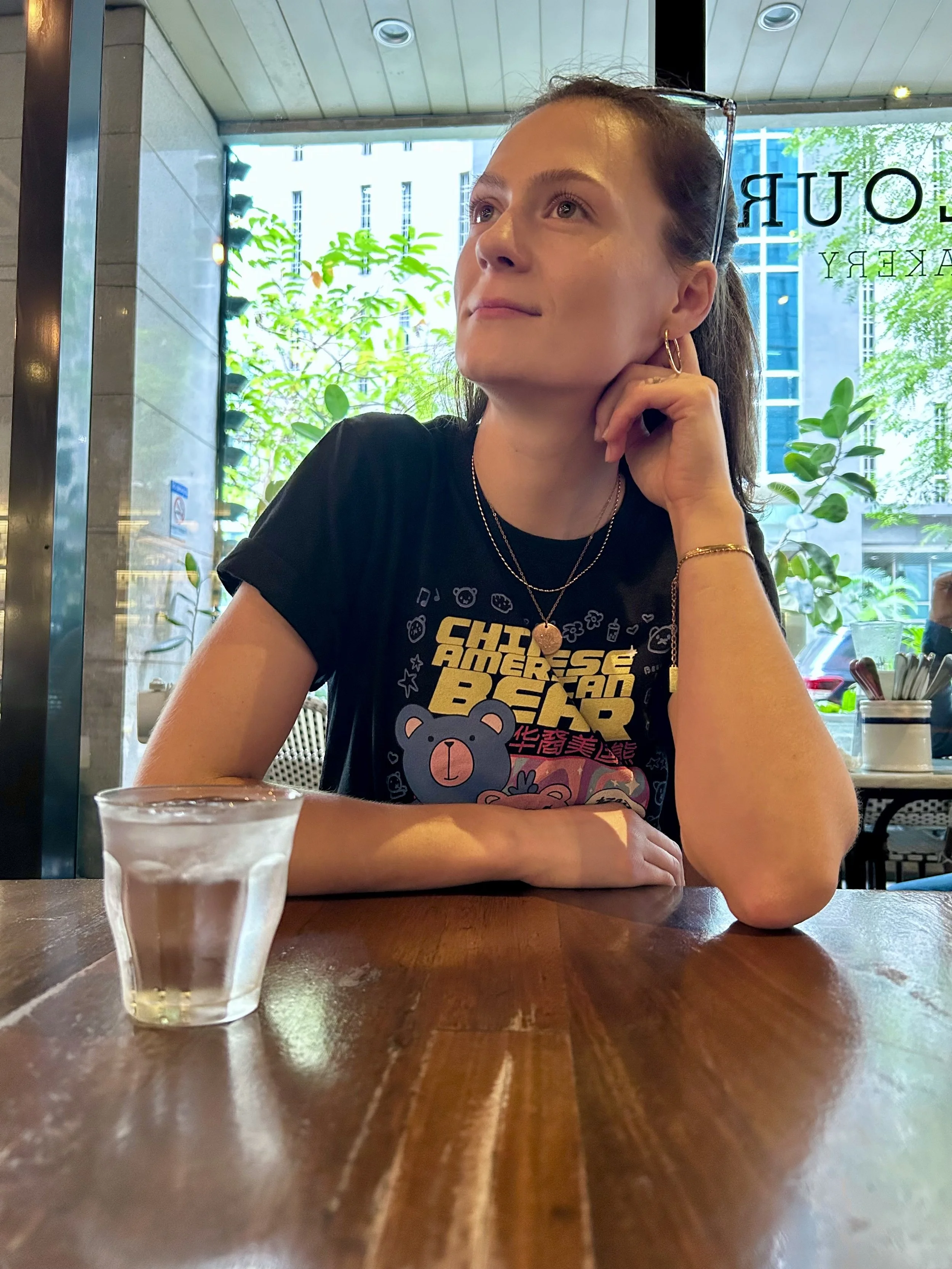 A woman sits at a table in a restaurant, with her hand resting on her chin, smiling and looking upwards. There is a shot glass with a clear liquid on the table in front of her, and large windows in the background showing buildings and trees outside.