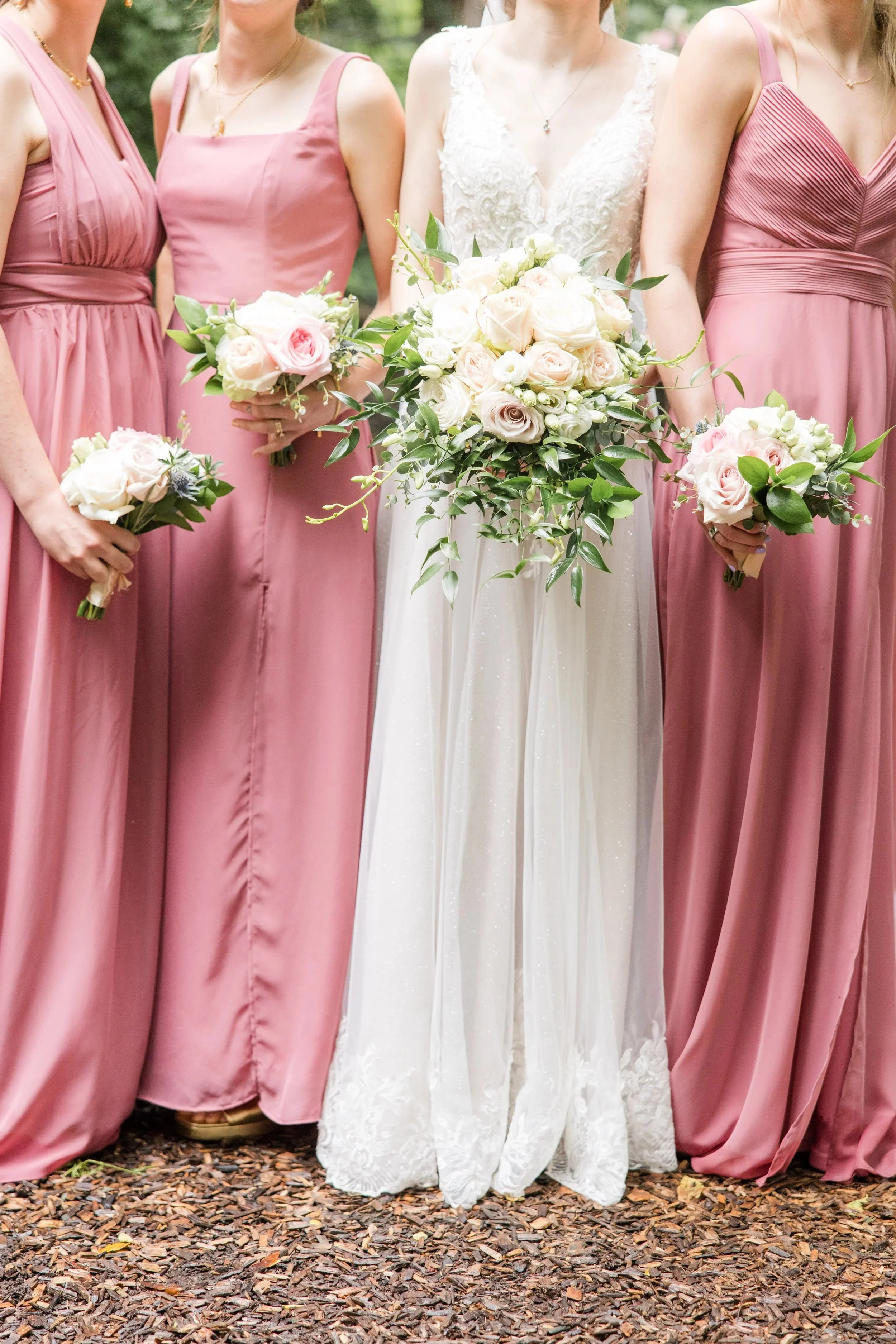 Four women at a wedding, with the bride in a white lace gown holding a large bouquet of white and pale pink roses, and three bridesmaids in matching pink dresses holding smaller bouquets of similar flowers, standing outdoors on a leaf-strewn ground.