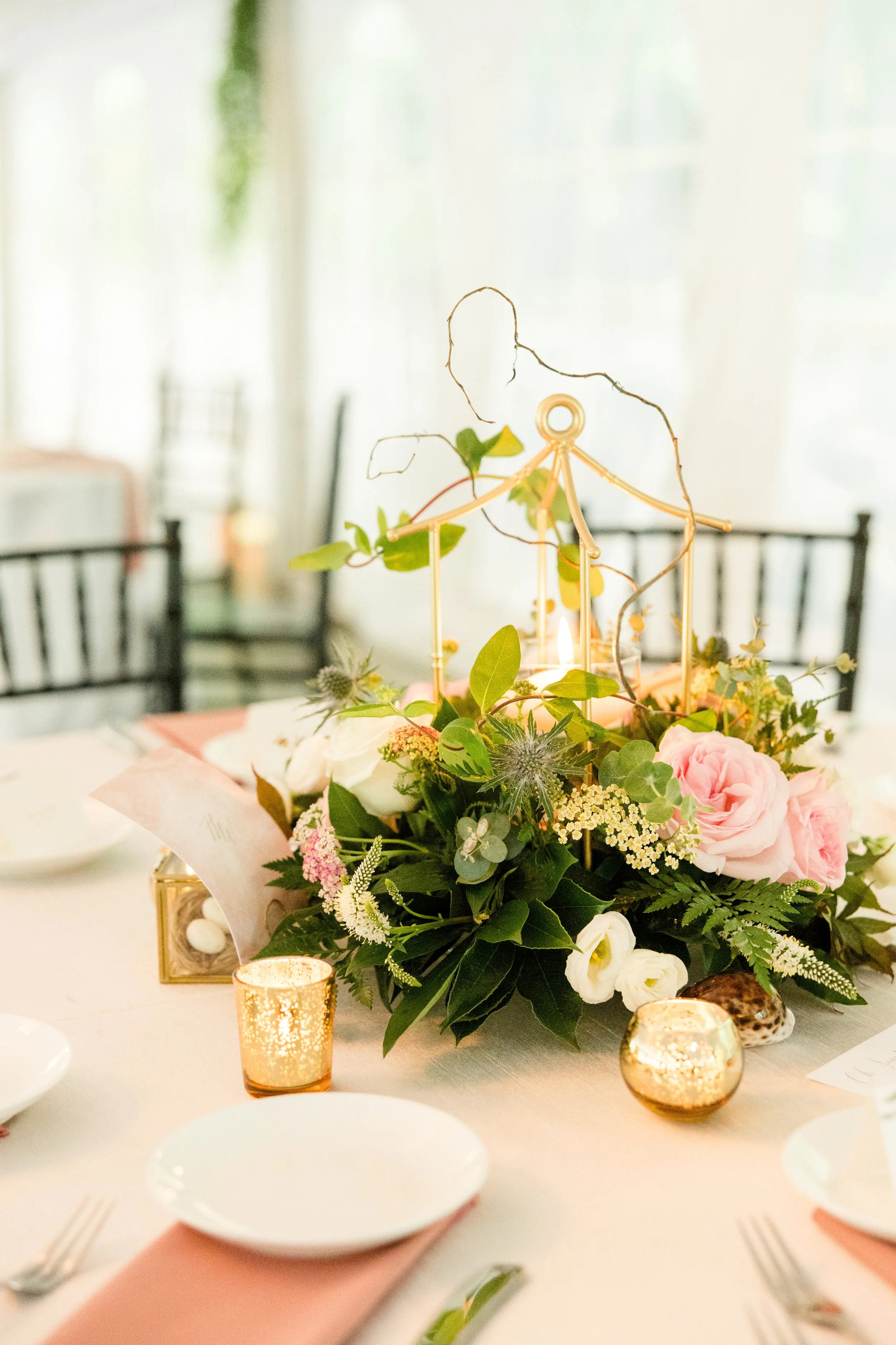 A floral centerpiece of pink, white, and purple flowers with greenery on a table. Surrounding the centerpiece are small candles in gold holders, place settings with white plates, and pink napkins. The background shows a bright, softly lit room with black chairs.