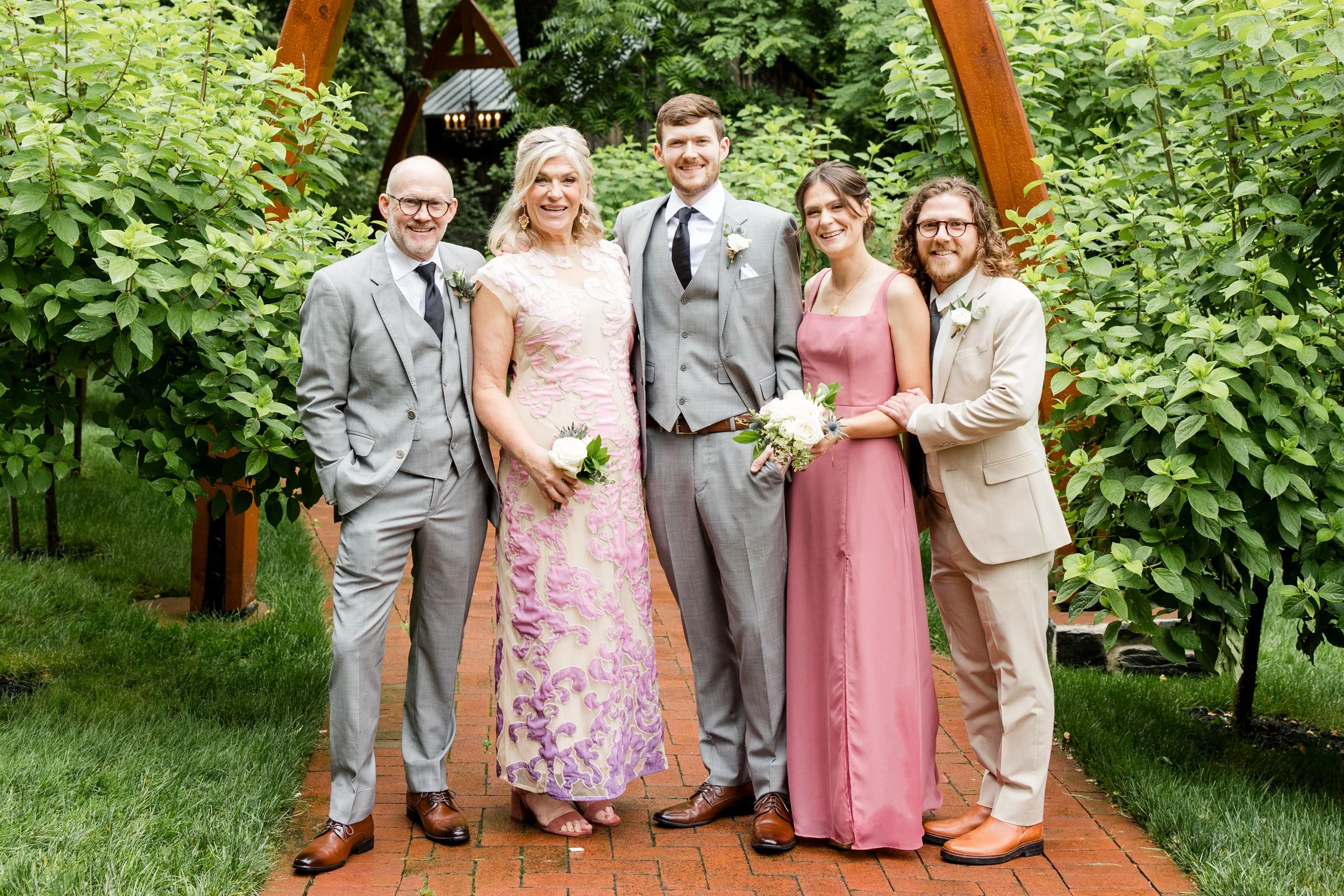 Group of six people dressed in formal attire, standing outdoors on a brick pathway with greenery and trees in the background, participating in a wedding celebration.