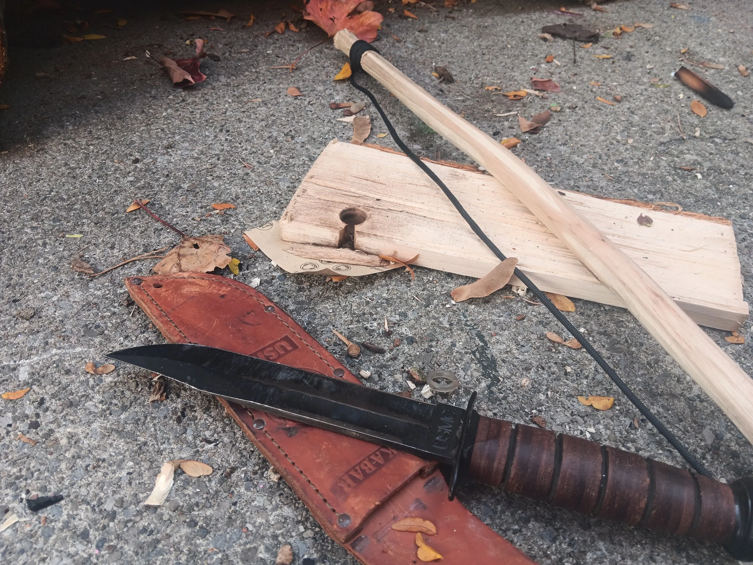 A black folding saw with a brown leather sheath on asphalt ground, surrounded by fallen leaves and pieces of wood.