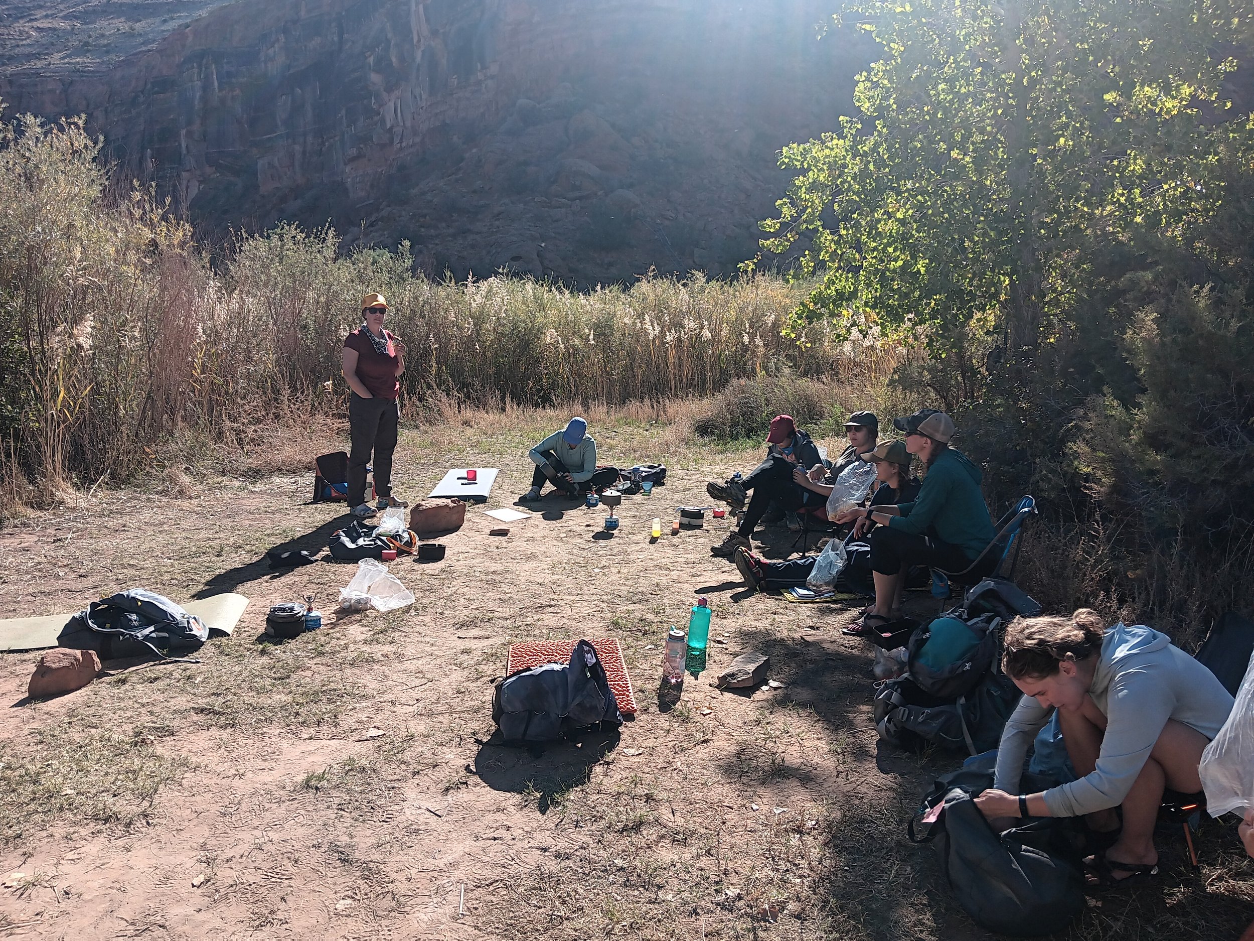 A group of people sitting outdoors on a dirt ground, surrounded by trees and rocky hills in the background. Some are sitting on the ground, some on chairs, with various backpacks, water bottles, and camping gear scattered around. One woman is standing and wearing sunglasses, a hat, and a scarf, holding a drink, while others are engaged in different activities like sitting and talking.
