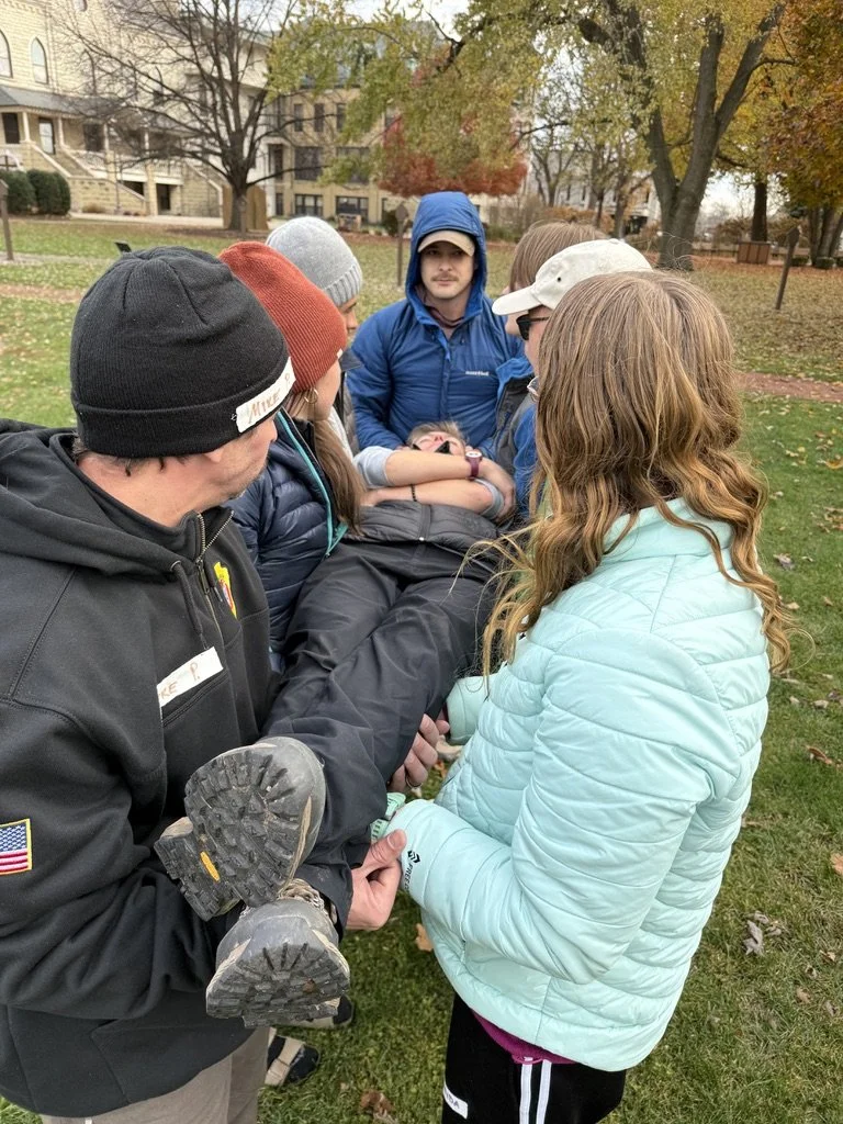 A group of people outdoors in a park, with some holding a person horizontally as if performing a rescue or drill. The scene includes trees with autumn foliage and residential buildings in the background.