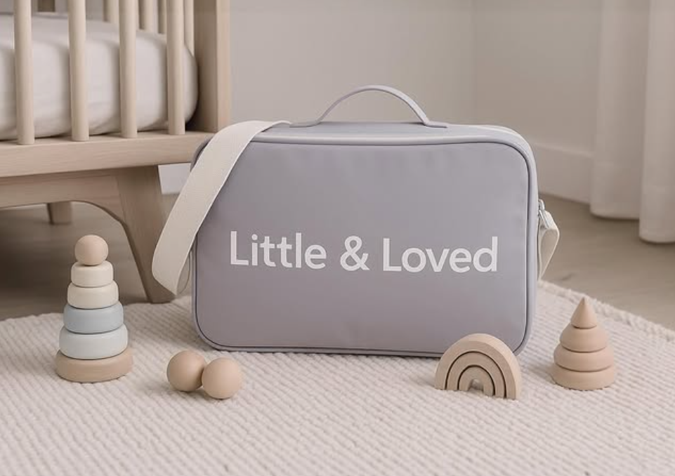 Una bolsa de transporte gris con la inscripción 'Little & Loved' en un entorno de habitación infantil, rodeada de juguetes de madera en forma de árbol, arcoíris y bolas pequeñas.