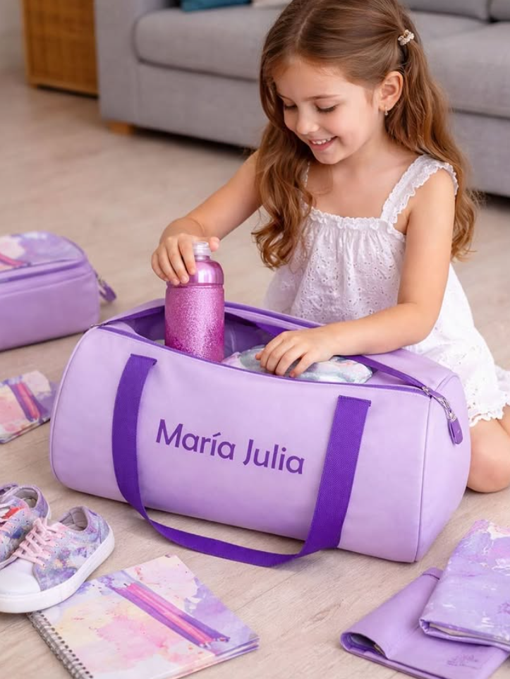 Niña sonriendo mientras prepara su bolso de viaje, que tiene el nombre 'María Julia', con botella rosa, zapatillas y materiales escolares alrededor.