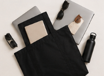 Bolsa de tela negra con objetos y accesorios sobre una superficie blanca, incluyendo una botella de agua, gafas de sol, una botella de spray y otros artículos pequeños.
