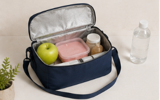 Estuche de almuerzo con una manzana verde, un recipiente rosa, un par de frascos (posiblemente para condimentos), y una botella de agua a un lado, sobre una mesa y una planta cerca.