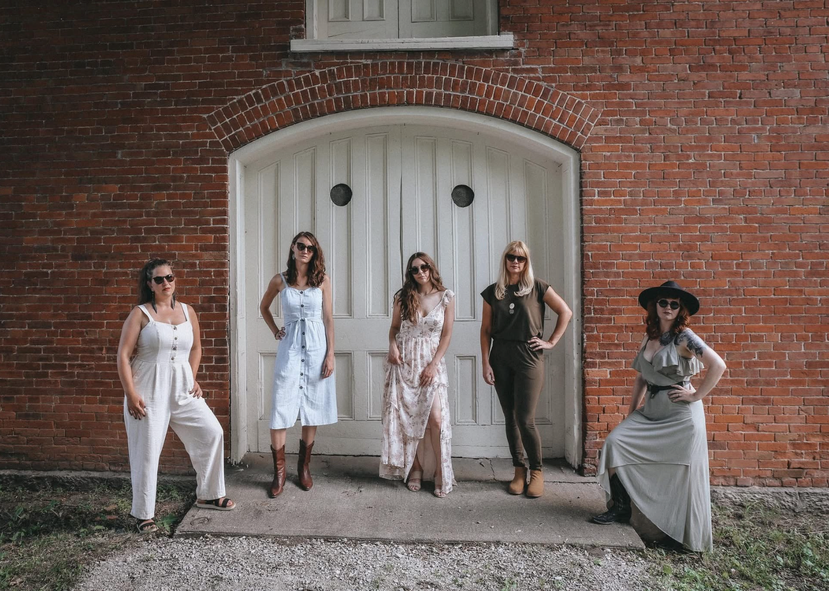Group of five women standing in front of a large white door set into a brick wall, posing for a photo. They are dressed in summer outfits and wearing sunglasses.