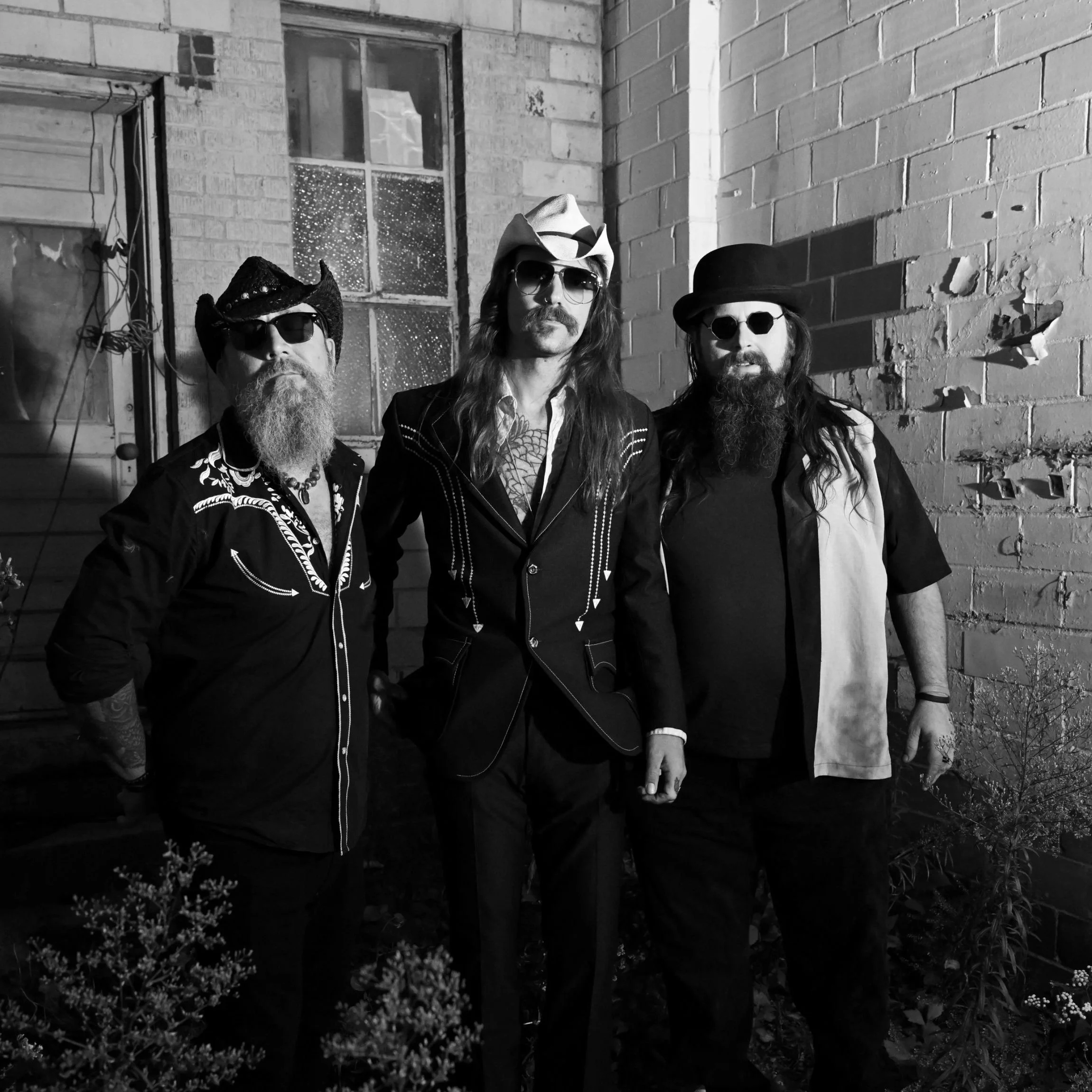Three men wearing sunglasses and hats standing against a brick wall under moonlight, with plants in foreground.