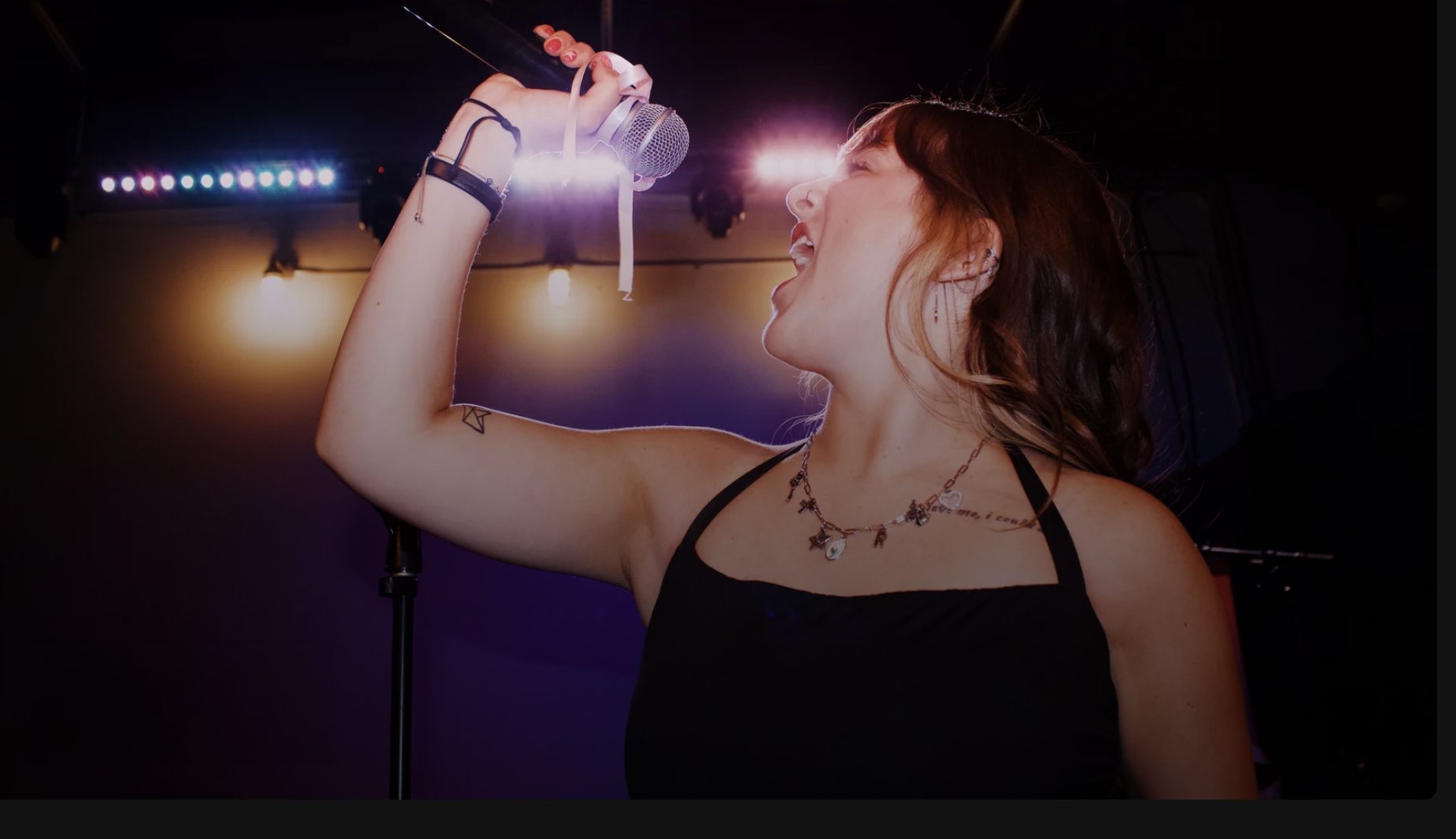 A woman singing into a microphone on stage, with colorful stage lights in the background, wearing a black tank top, layered necklaces, and tattoos on her arm and chest.