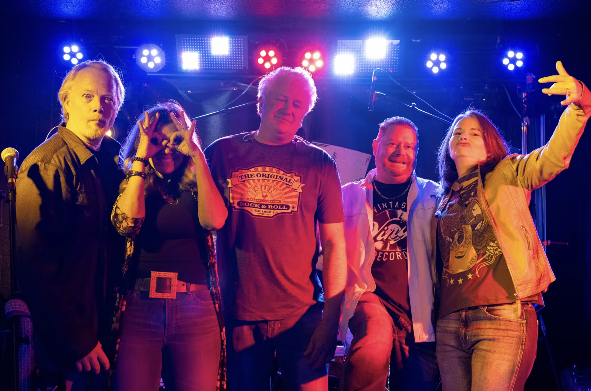 A group of five people on stage at a concert, under colorful stage lights, smiling and making playful gestures.