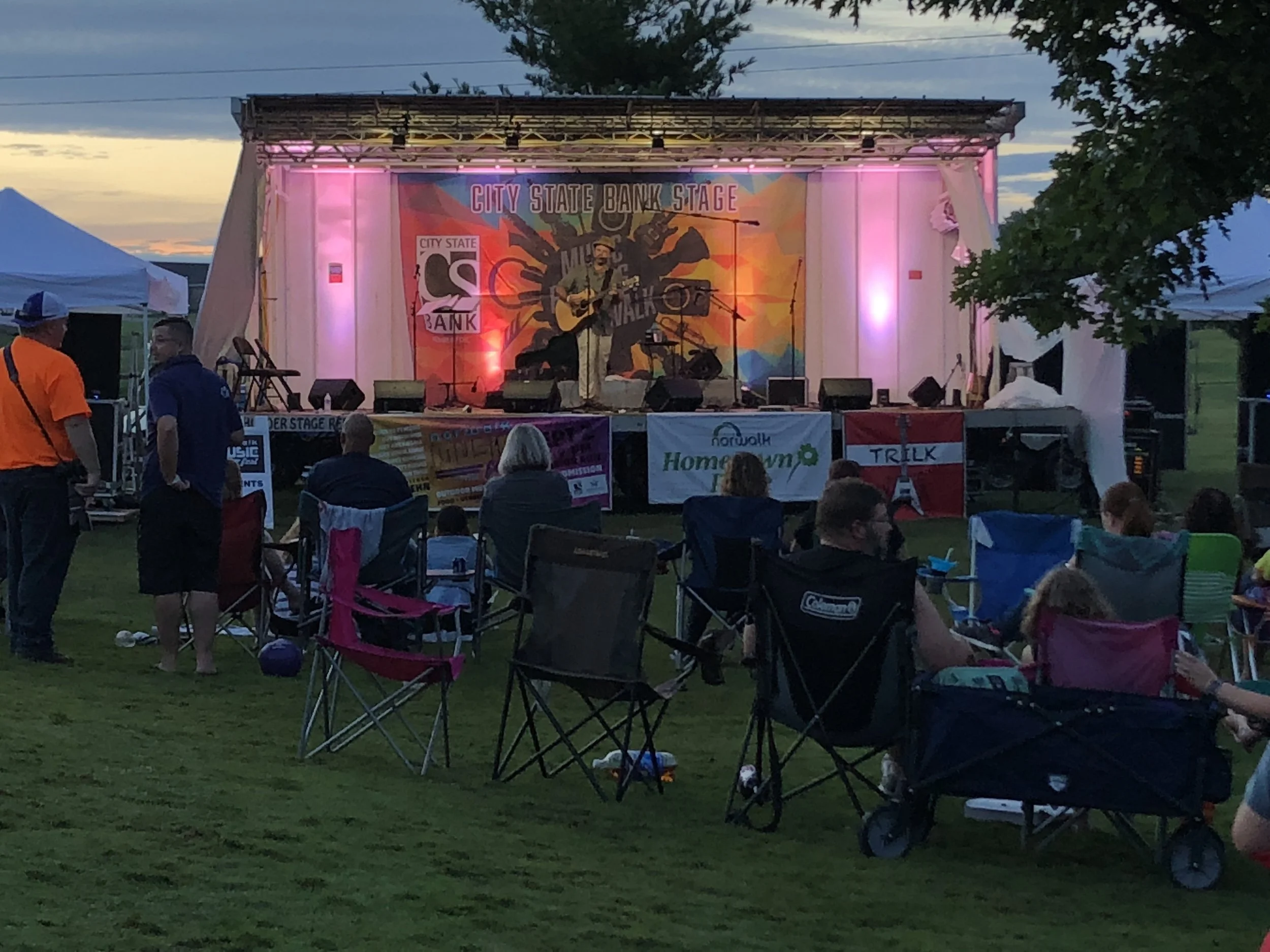 Outdoor concert stage with a man playing guitar and singing, surrounded by audience seated in chairs at dusk, trees and tents visible around the stage.