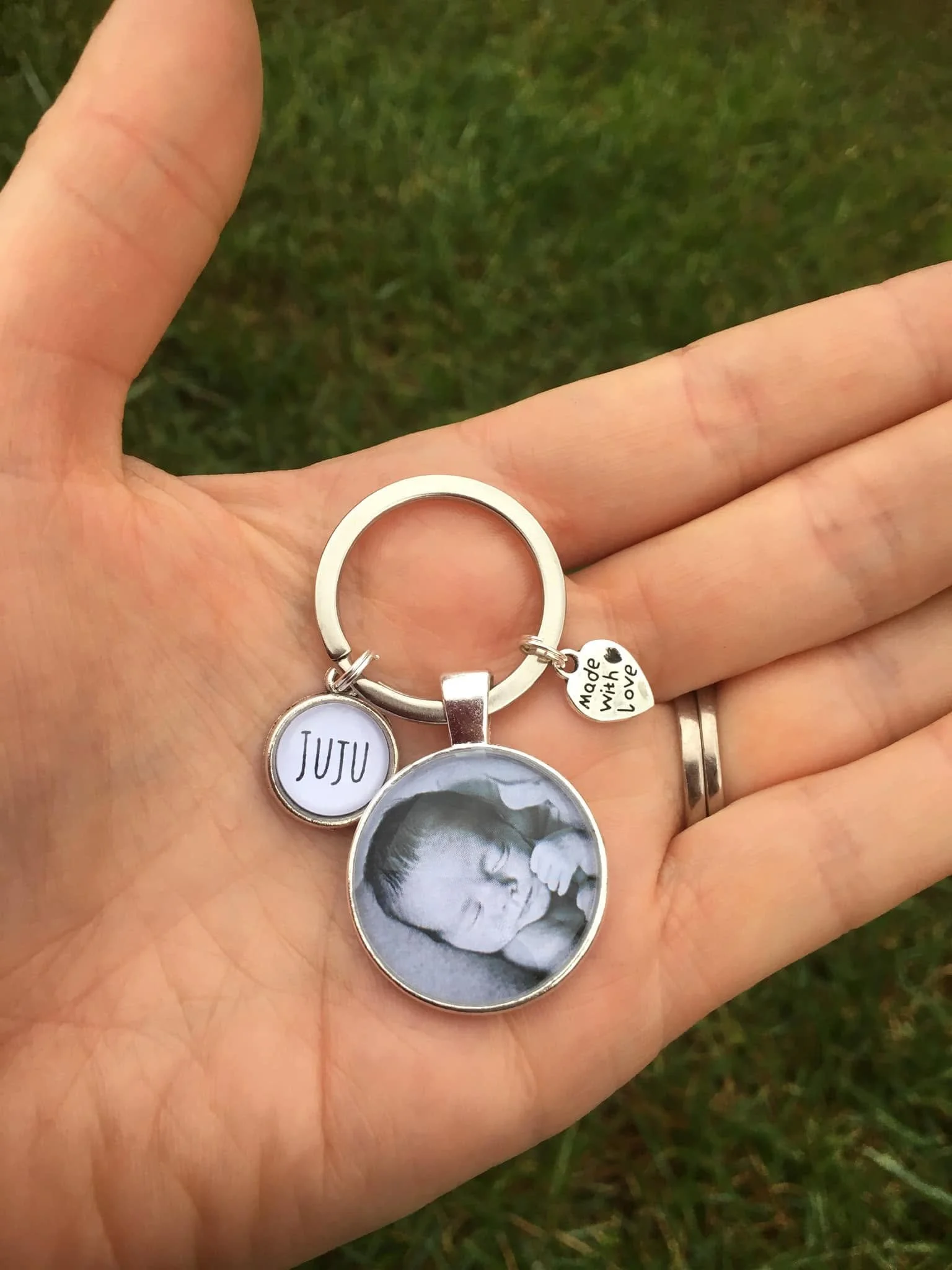 A hand holds a keychain with several charms, including a circular photo of a sleeping baby, a white circle with the name "JUJU", and a small heart-shaped charm engraved with "Made with Love". The background is grass.