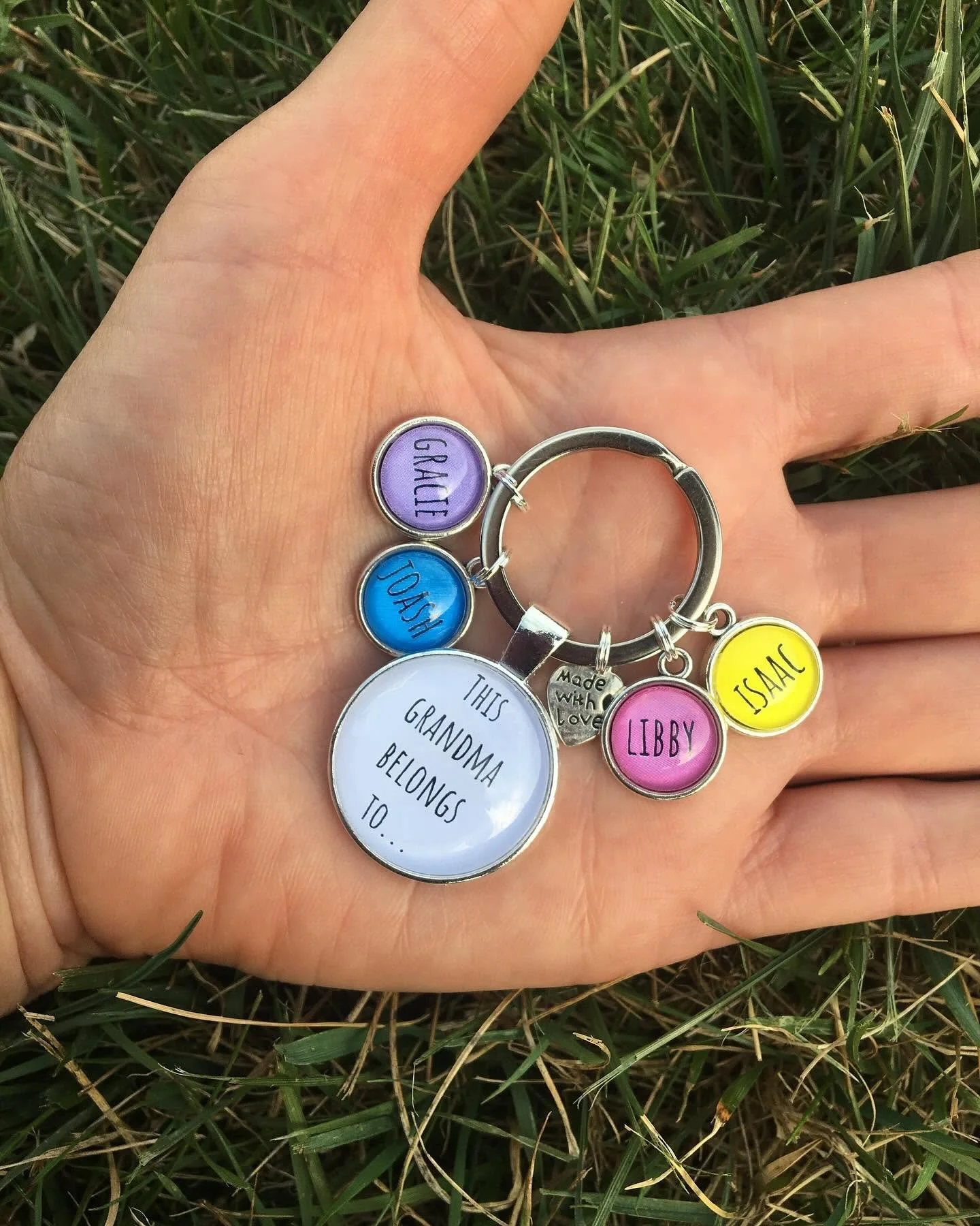 A hand holds a keychain with several colorful charms, including one purple with 'GRACE,' one blue with 'JOSH,' one yellow with 'ISAAC,' a pink one with 'LIBBY,' and a round white one with the text 'THIS GRANDMA BELONGS TO...' in black, all lying on green grass.