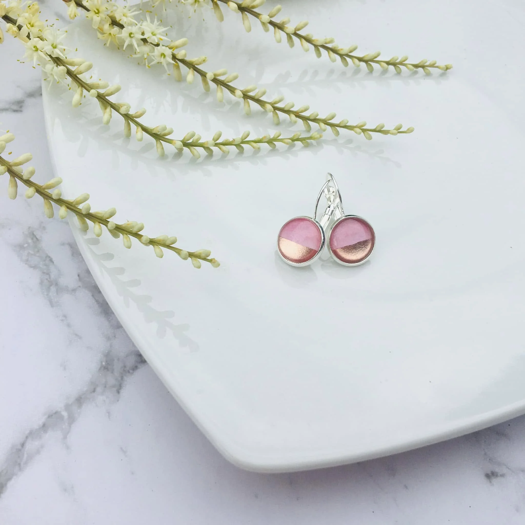 Close-up of cream-colored flowers with long stems and small buds on a white surface with a marble pattern, with pink earrings with metallic accents at the bottom.