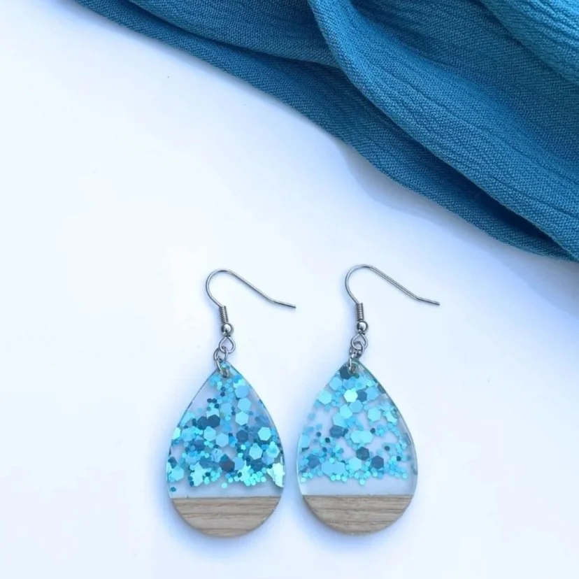 Pair of teardrop-shaped earrings with blue glitter and wood accents on a white background, with a blue fabric in the corner.