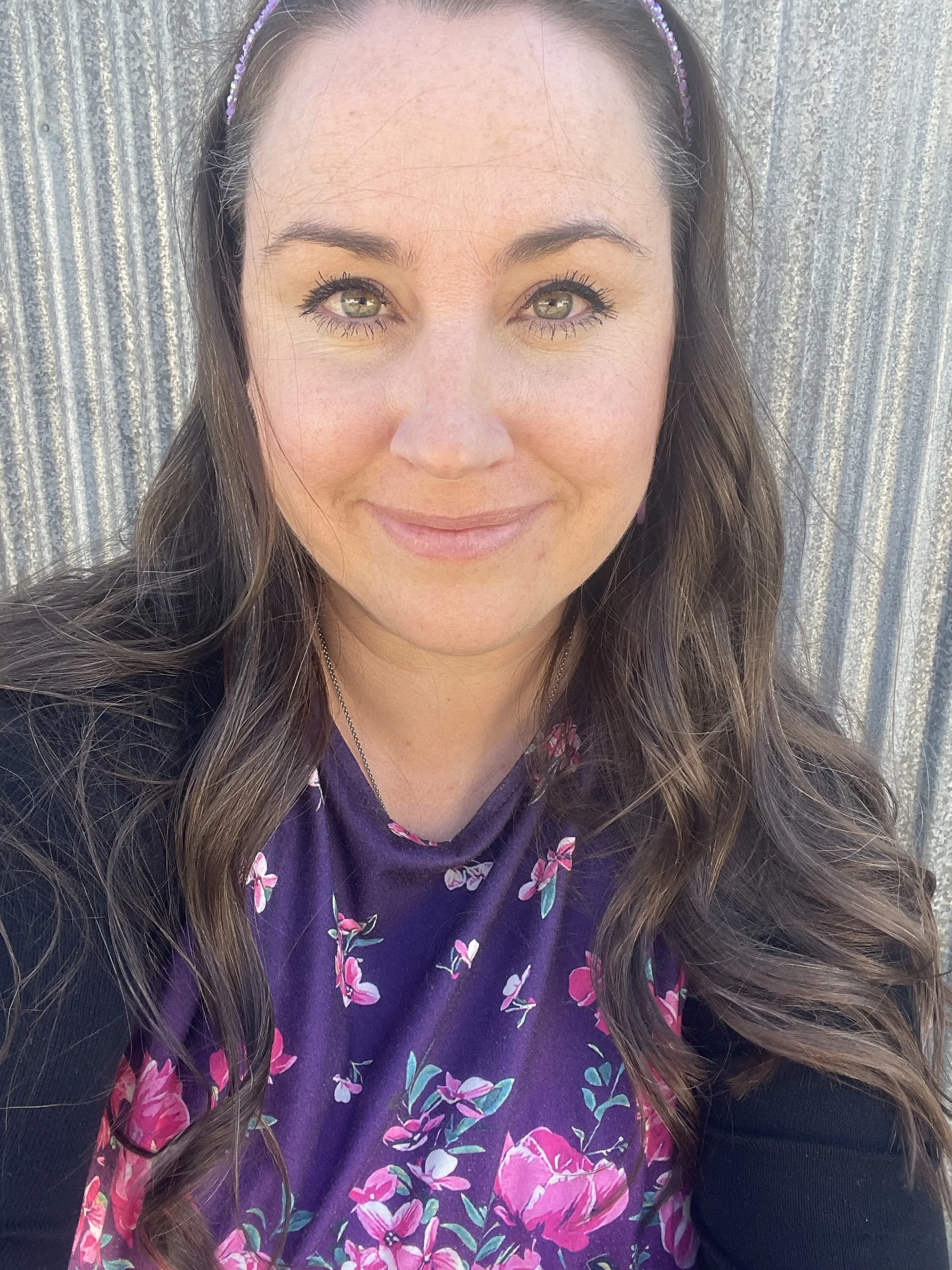 Close-up of a woman with long brown hair, green eyes, light makeup, wearing a purple headband and a purple dress with a pink and white floral pattern, standing outside in front of a weathered metal wall.