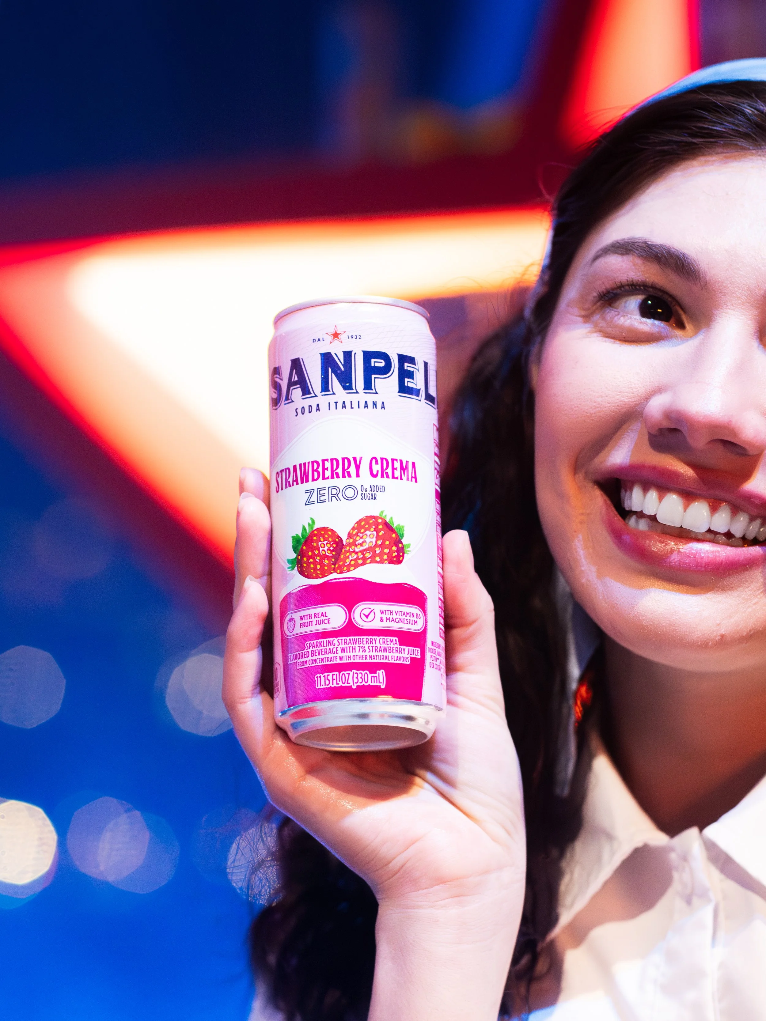 A young woman smiling and holding a can of San Pellegrino Strawberry Crema soda close to her face. The background is colorful with bokeh effects.