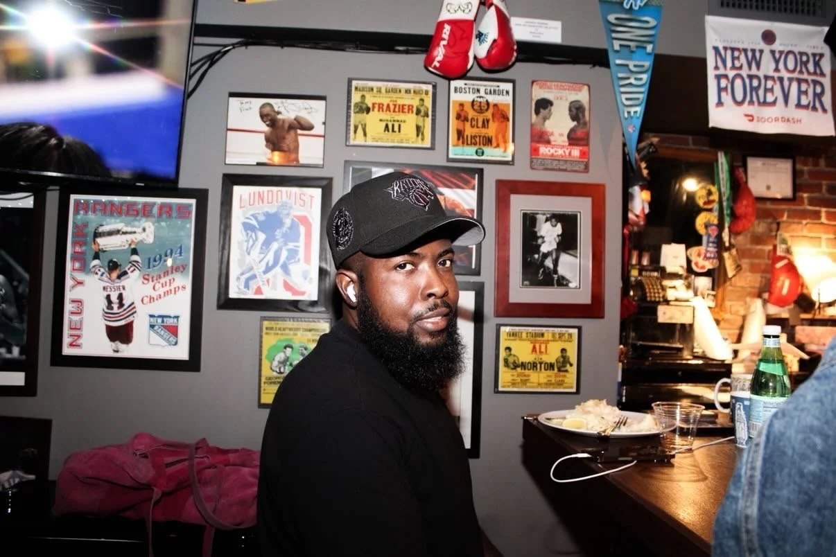 A man with a beard and wearing a black cap and shirt sitting at a table in a sports-themed restaurant or bar decorated with sports posters, photographs, and memorabilia on the wall.